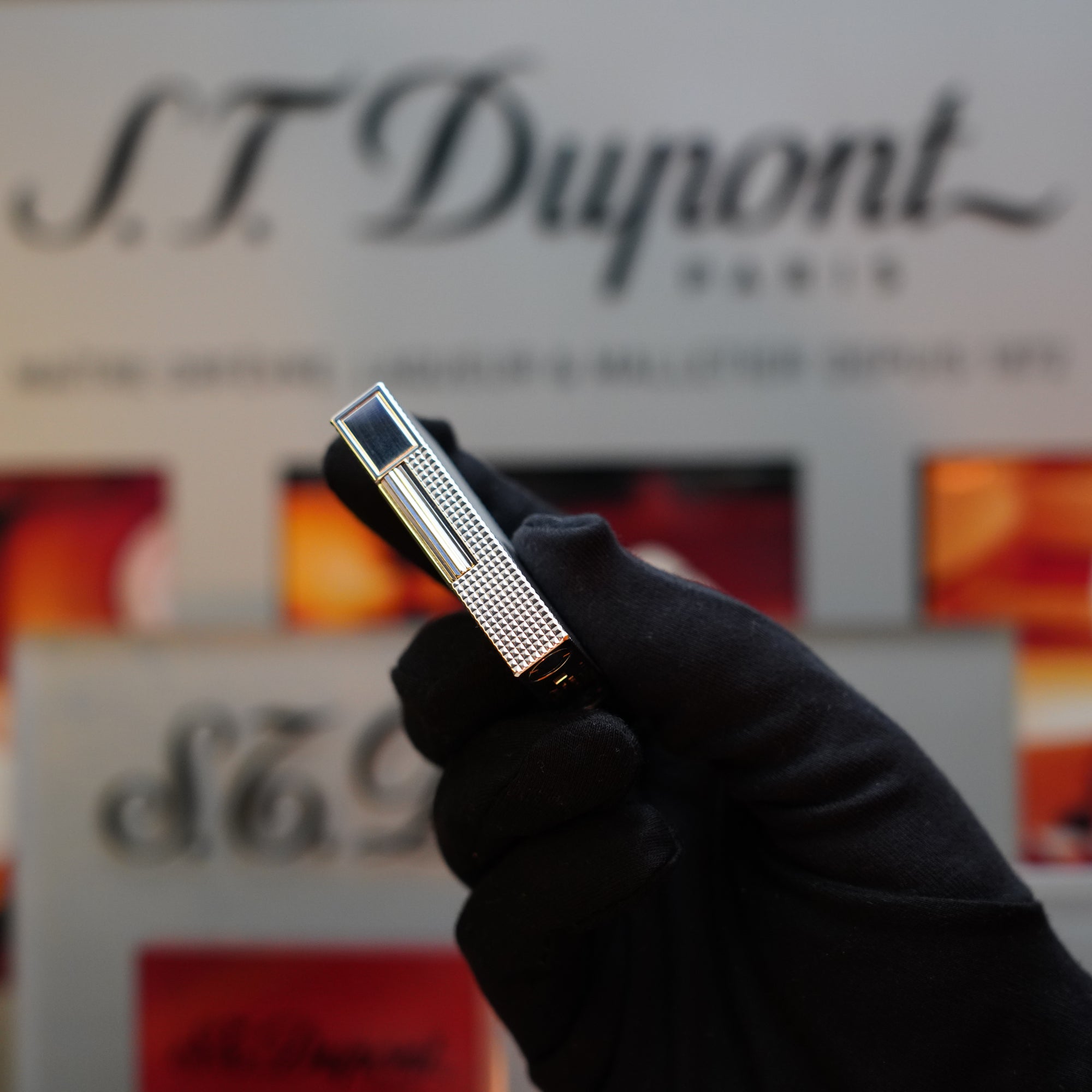 A gloved hand holds a 1970 S.T. Dupont Diamond Head Lighter Heavy Silver Finish ligne 2, its silver finish gleaming, with the company logo blurred in the background.