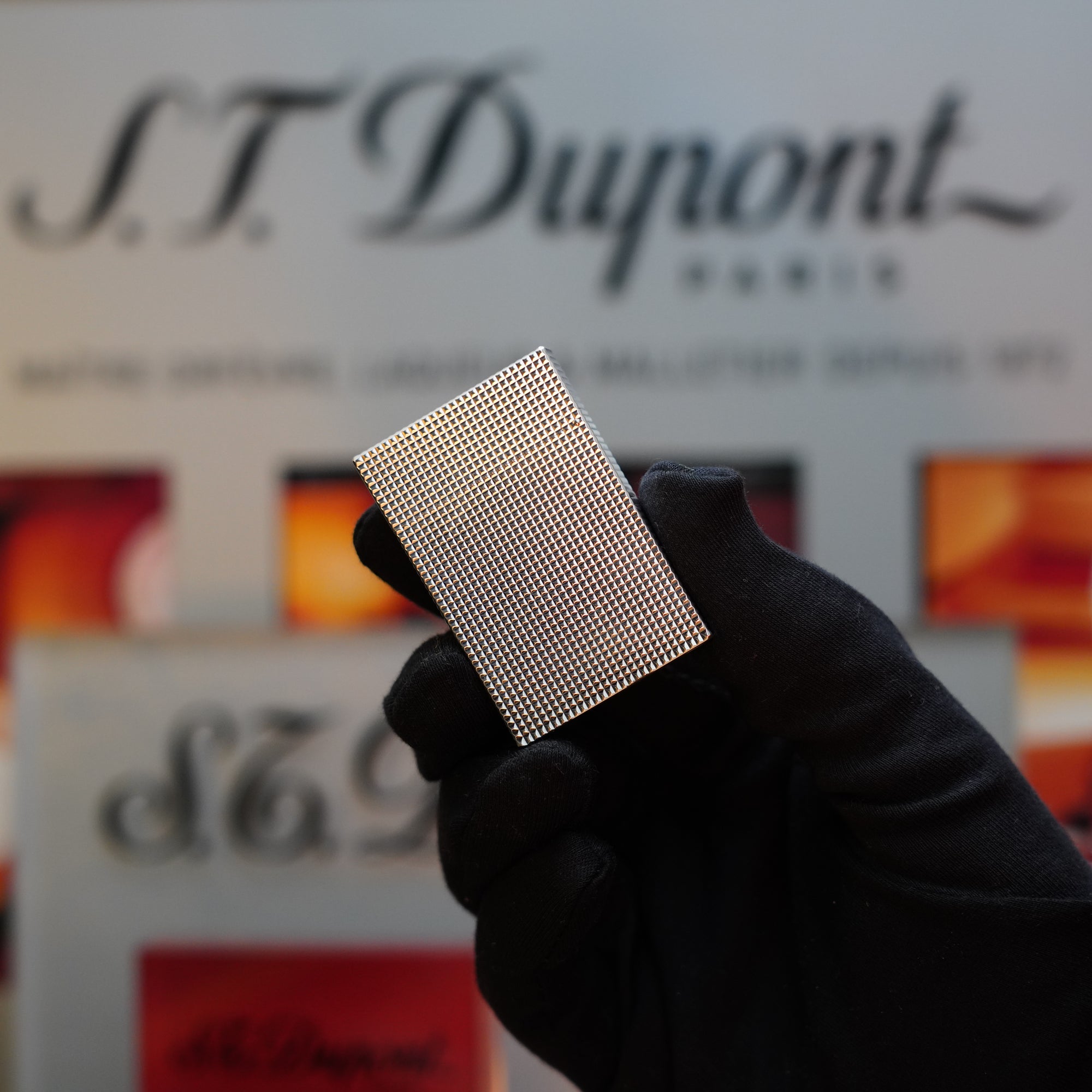 A gloved hand holds a metallic, textured rectangular object in front of an S.T. Dupont Paris sign—an exquisite 1970 St Dupont Diamond Head Lighter Heavy Silver Finish ligne 2.