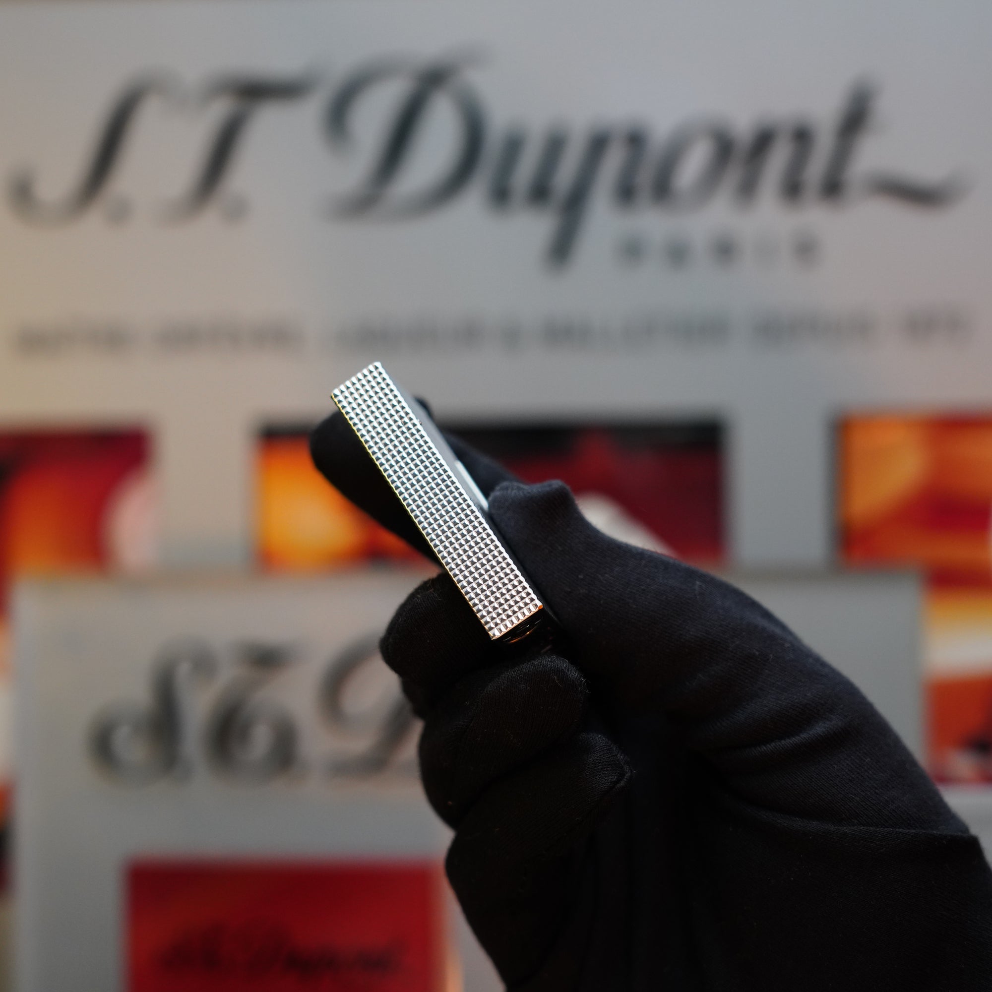 A gloved hand holds a 1970 St Dupont Diamond Head Lighter Heavy Silver Finish ligne 2 in front of a blurred background with 'S.T. Dupont' branding, reminiscent of the elegant Ligne 2 Series.