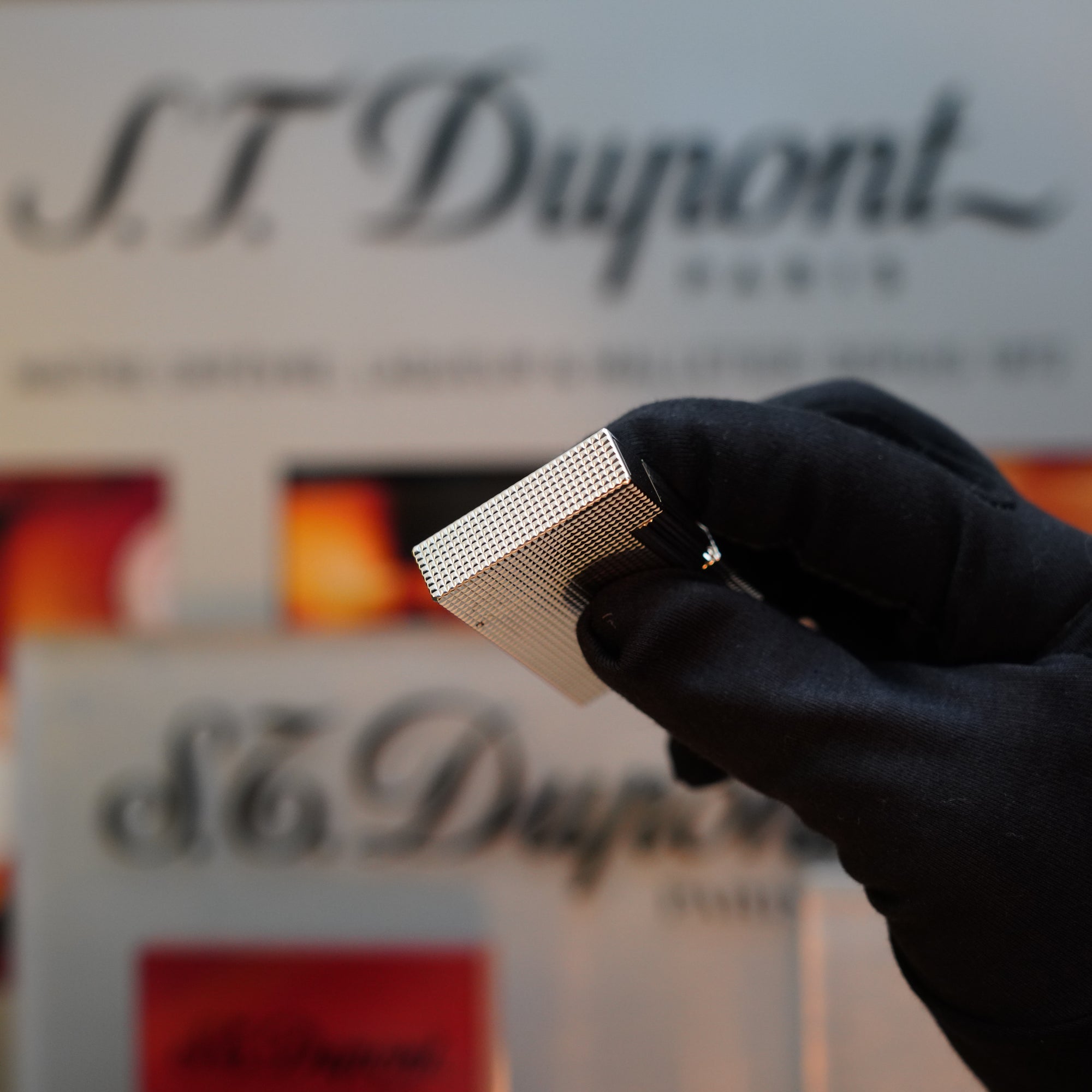 A gloved hand holds a 1970 St Dupont Diamond Head Lighter Heavy Silver Finish from the Ligne 2 series, showcasing its silver-colored grid pattern against a blurred background displaying the S.T. Dupont logo.