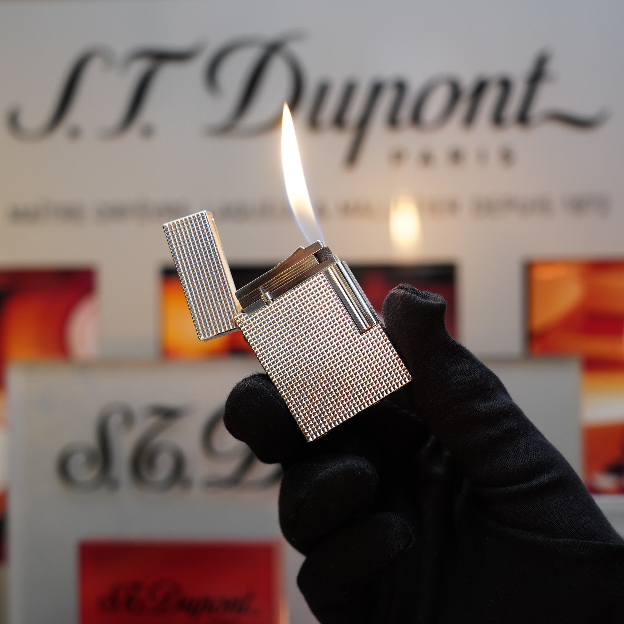 A person wearing a black glove holds a lit, silver, textured 1970 St Dupont Diamond Head Lighter Heavy Silver Finish Ligne 2 in front of an S.T. Dupont Paris sign.