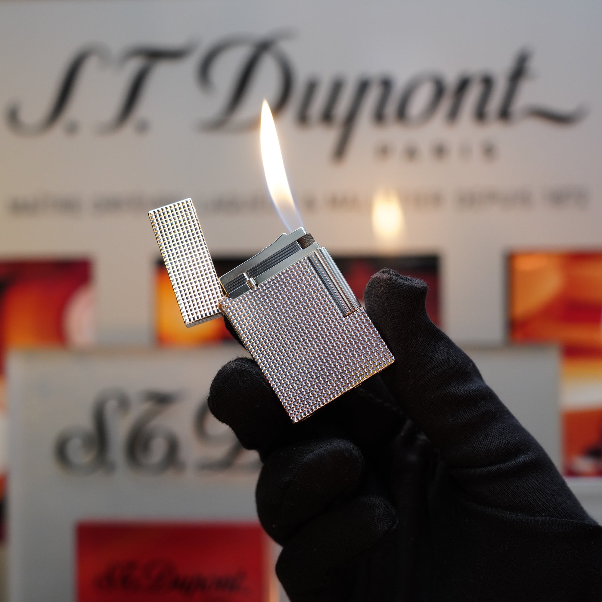 A hand in a black glove is igniting a 1970 St Dupont Diamond Head Lighter Heavy Silver Finish from the Ligne 2 Series. The background features the iconic S.T. Dupont logo and other blurred signs, highlighting the timeless elegance of this vintage S.T. Dupont lighter.