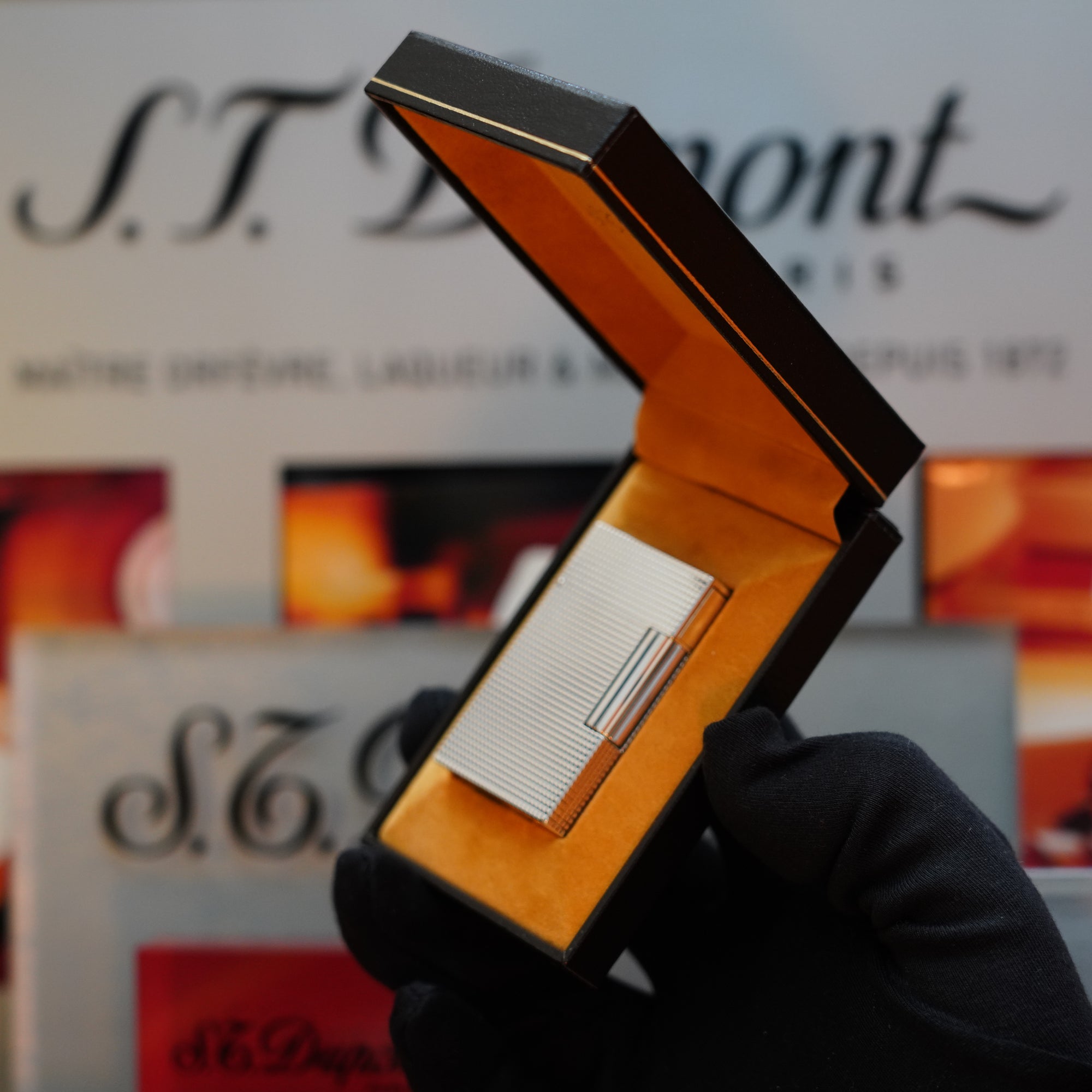 A gloved hand holds an open box with a 1970 St Dupont Diamond Head Lighter Heavy Silver Finish ligne 2 inside, set against a blurred background featuring S.T. Dupont branding.