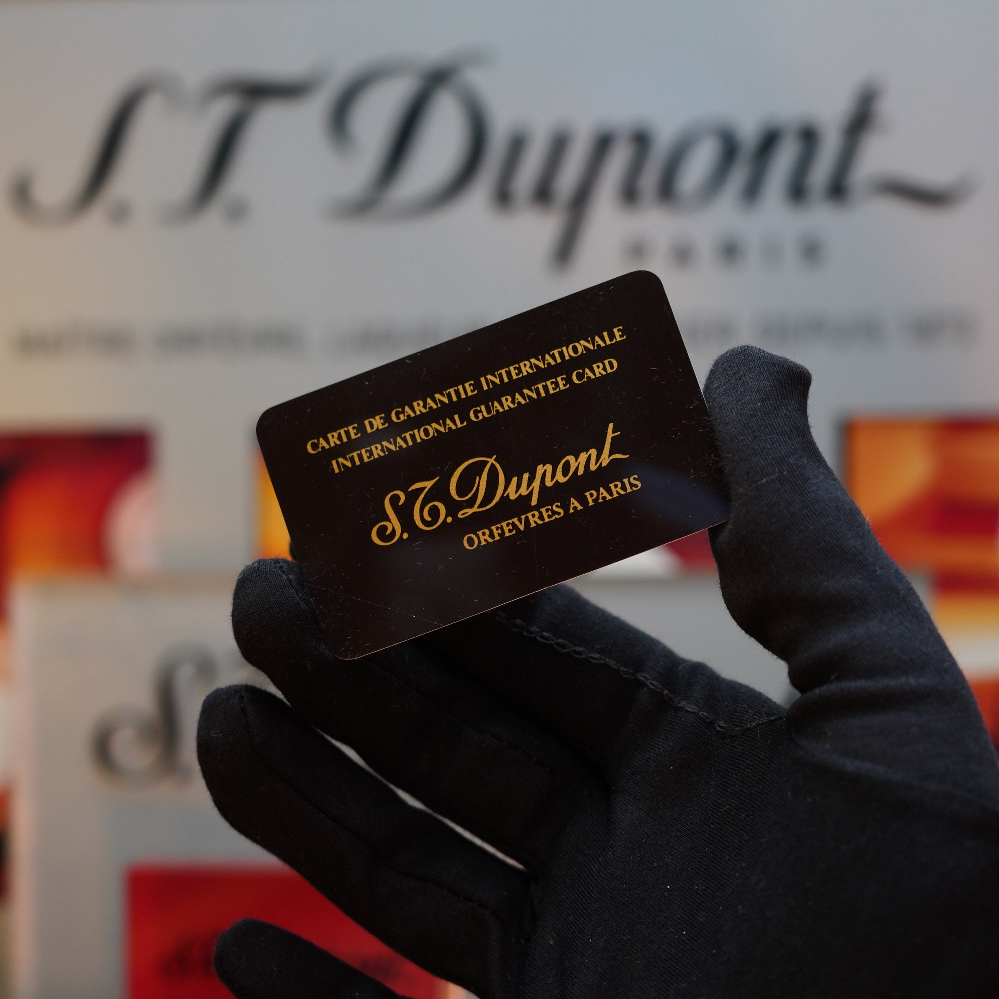 A gloved hand holds a black S.T. Dupont International Guarantee Card with gold text. In the blurred background, the iconic S.T. Dupont branding appeals, reflecting the timeless elegance of the 1970 St Dupont Diamond Head Lighter Heavy Silver Finish Ligne 2 Series and its vintage allure.