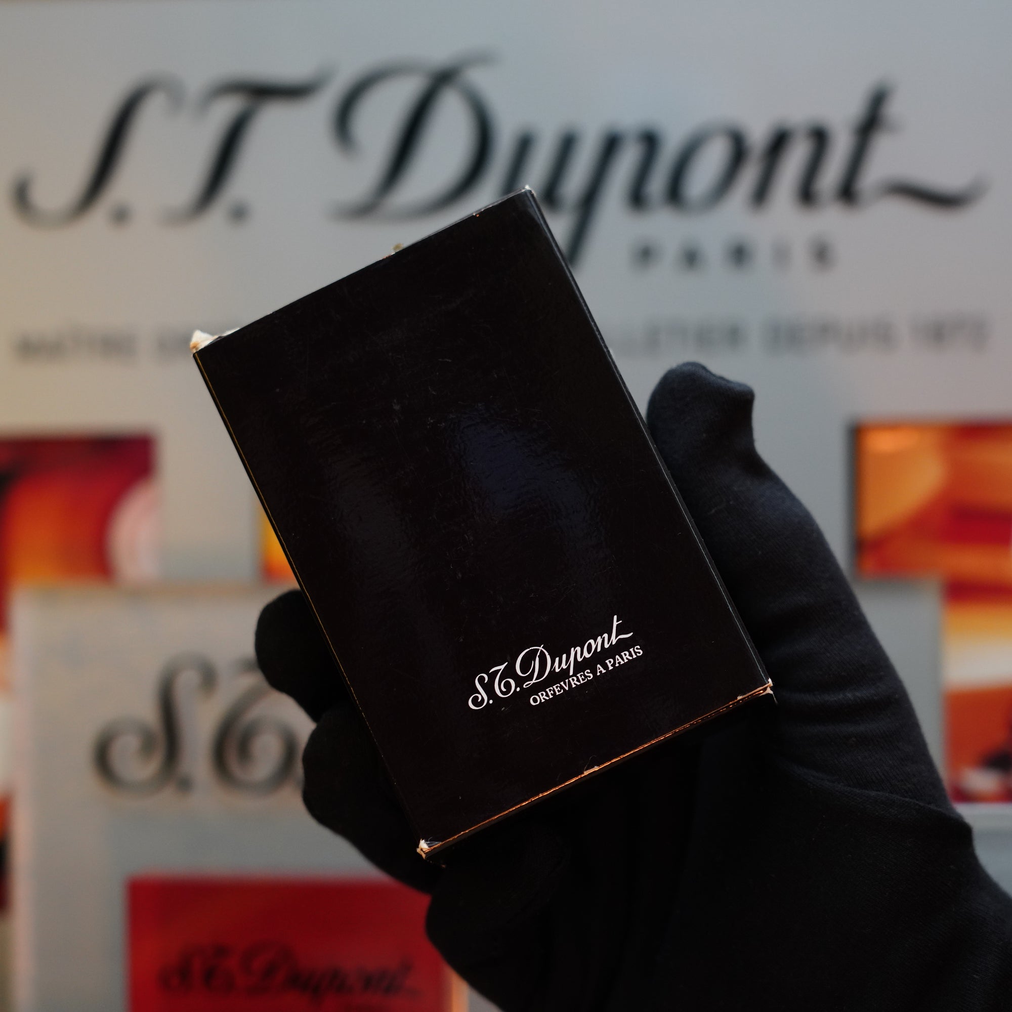 A gloved hand holds a black box branded with the S.T. Dupont logo, hinting at the elegance of the 1970 S.T. Dupont Diamond Head Lighter Heavy Silver Finish Ligne 2 series. In the blurred background, more S.T. Dupont branding whispers tales of timeless sophistication.