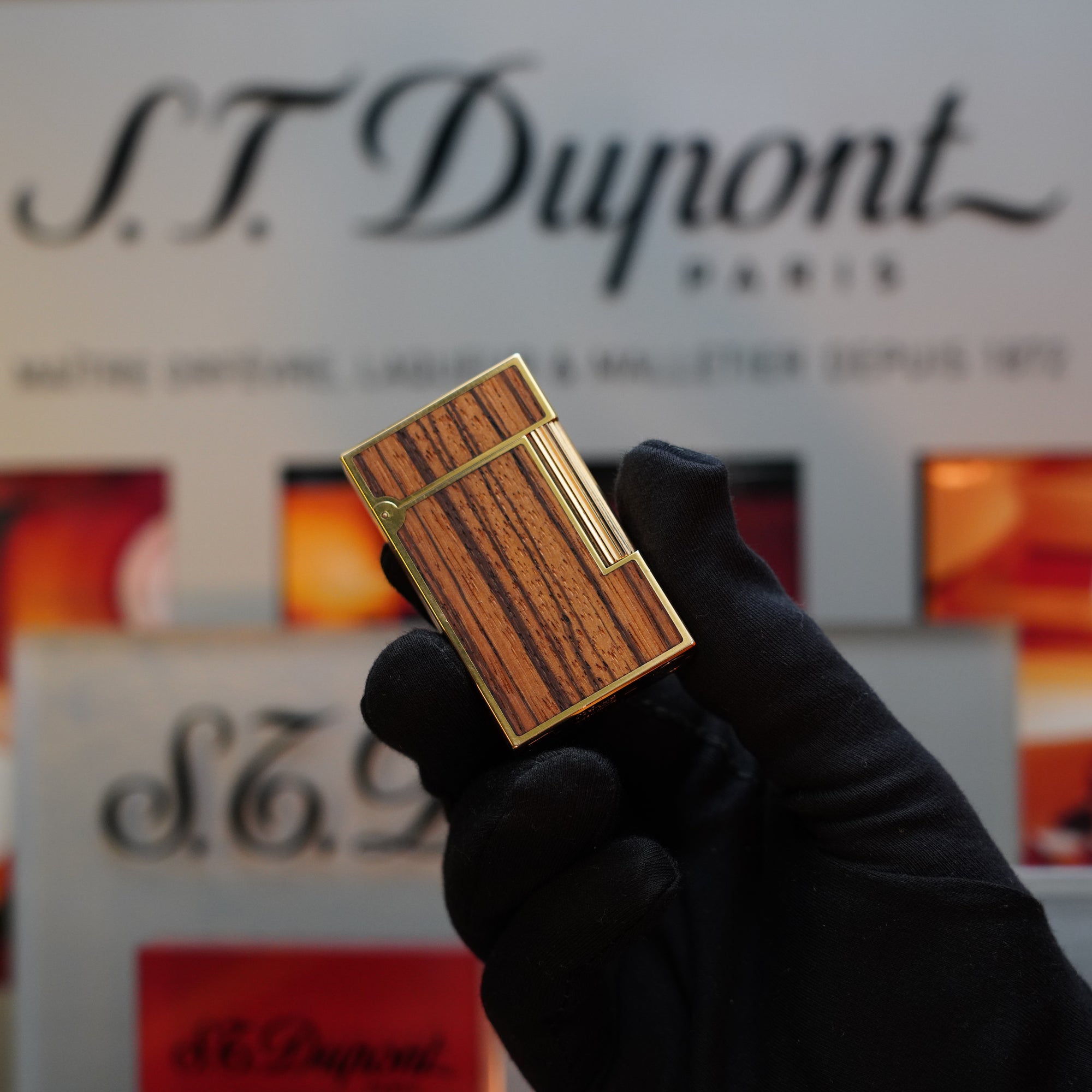 A gloved hand holds a 1989 S.T. Dupont Real Wood no lacquer, prototype, extremely rare Ligne 2 lighter in front of a blurred S.T. Dupont Paris sign—a true collector's item.