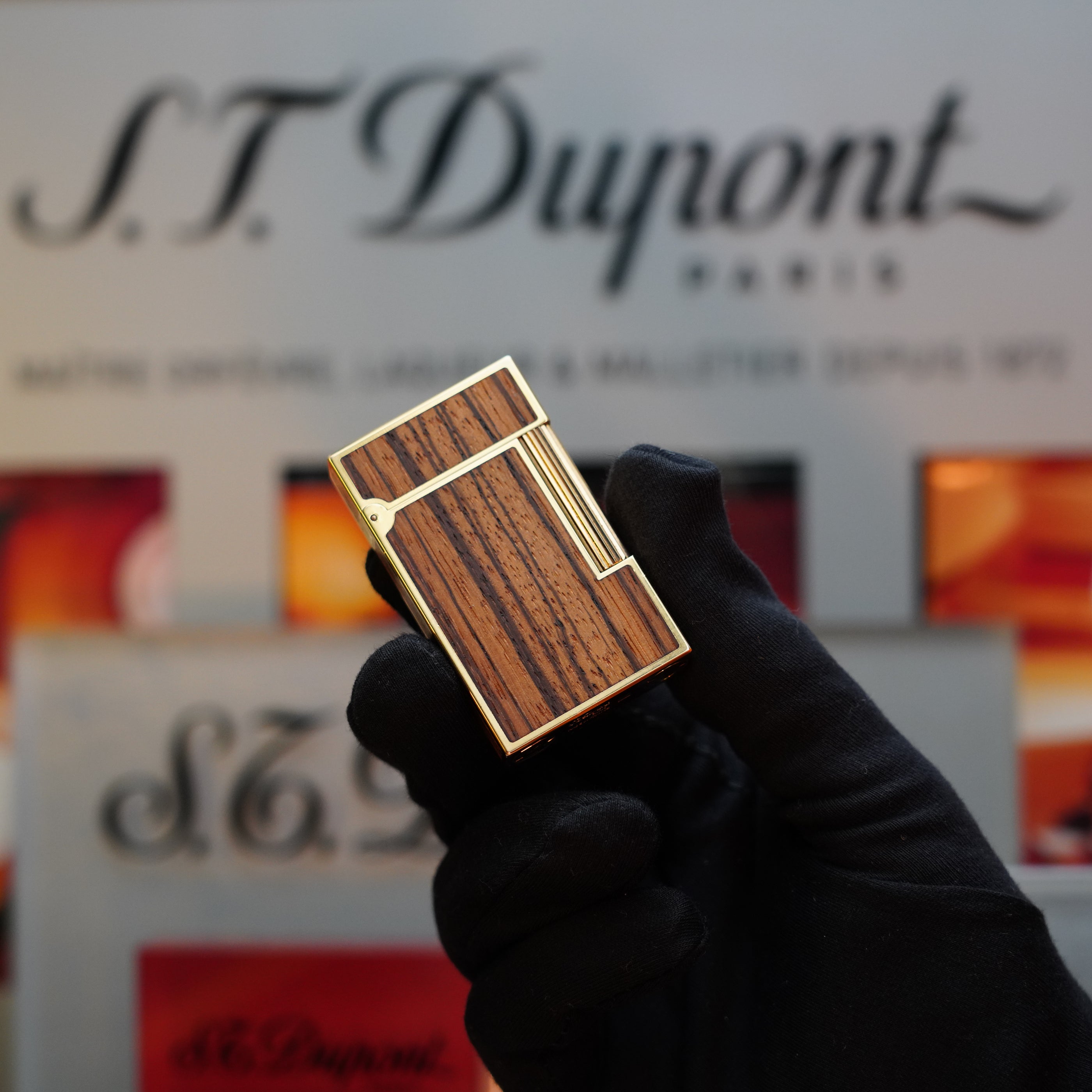 A gloved hand holds a luxury 1989 S.T. Dupont Real Wood no lacquer, prototype, extremely rare Ligne 2 lighter in front of a blurred S.T. Dupont Paris logo background.