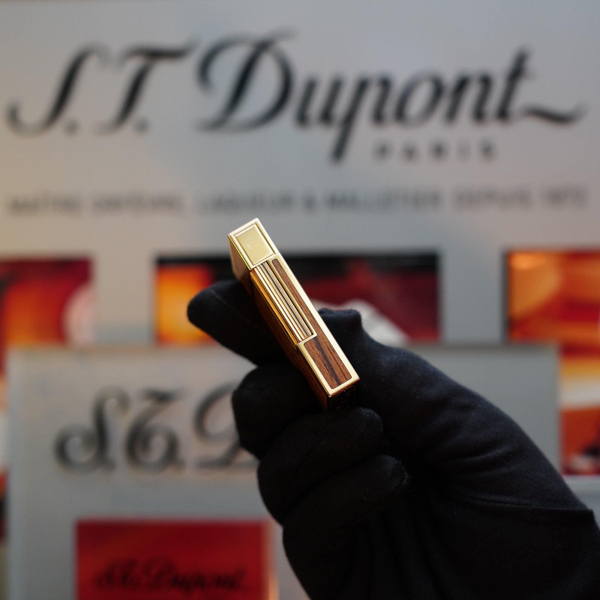 A gloved hand holding a 1989 S.T. Dupont Real Wood no lacquer, prototype, extremely rare Ligne 2 lighter in front of a blurred S.T. Dupont Paris sign.