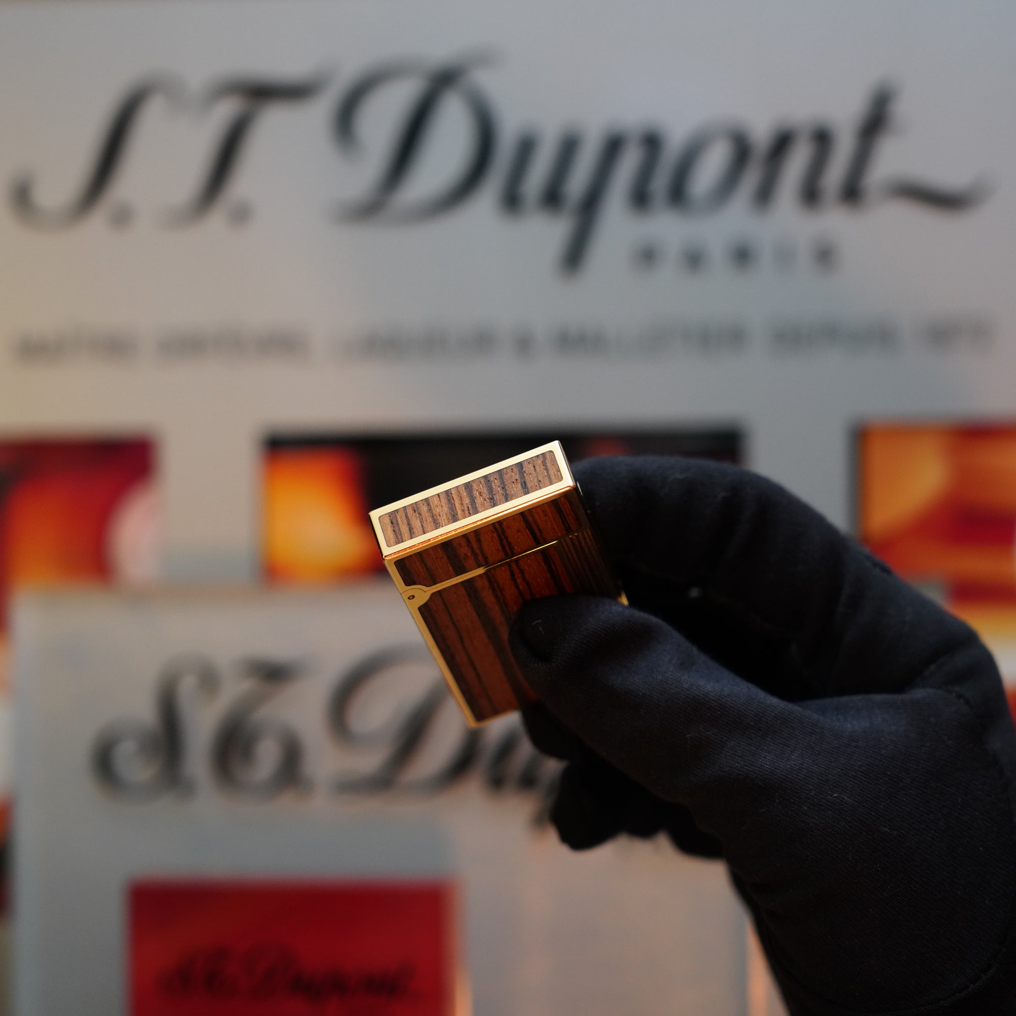 A gloved hand holds a 1989 S.T. Dupont Real Wood No Lacquer prototype Ligne 2 lighter in front of a blurred background featuring S.T. Dupont branding and various product images, showcasing an extremely rare collector's item from their Limited Prototype Series.