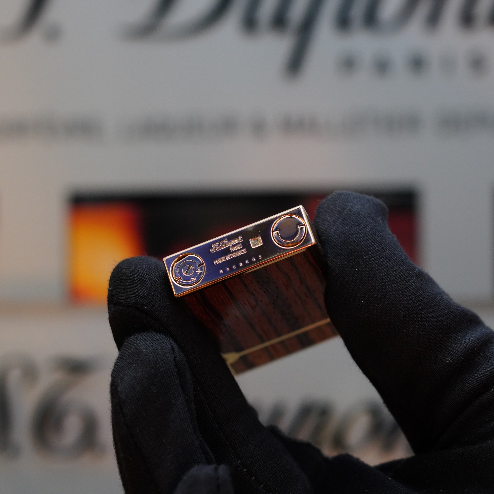 A gloved hand holds a small rectangular object with a metallic end featuring logos and text, reminiscent of a vintage lighter, against a blurred background. The object is identified as the 1989 S.T. Dupont Real Wood no lacquer, prototype, extremely rare Ligne 2 lighter.