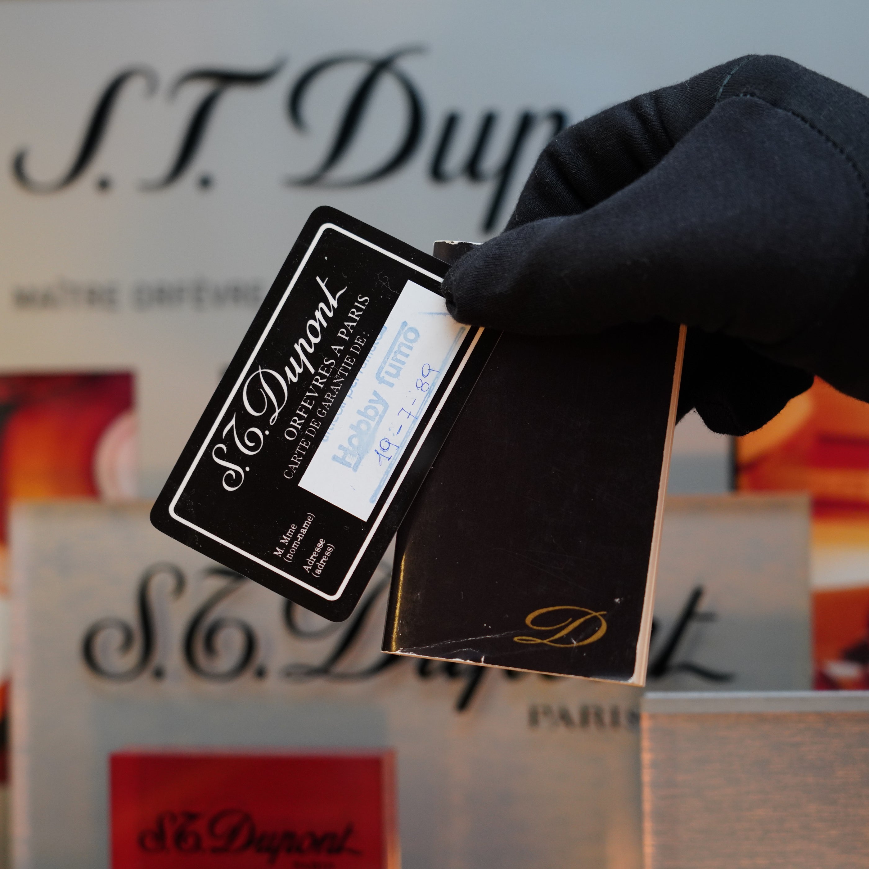 A gloved hand holds a vintage S.T. Dupont certificate card dated 1989, surrounded by various S.T. Dupont branded items including the extremely rare, no lacquer, real wood Ligne 2 lighter from the Limited Prototype Series.