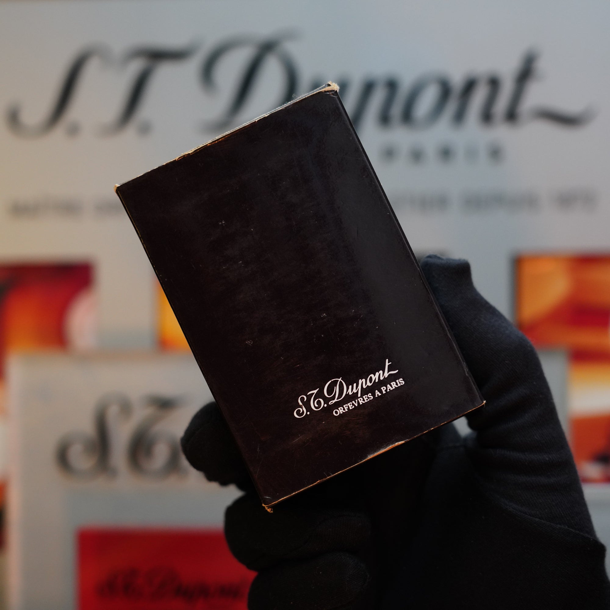 A gloved hand holds a black S.T. Dupont box, showcasing a 1989 Real Wood no lacquer prototype lighter from the extremely rare Ligne 2 series, against a blurred background with S.T. Dupont branding, making it a true collector's item.