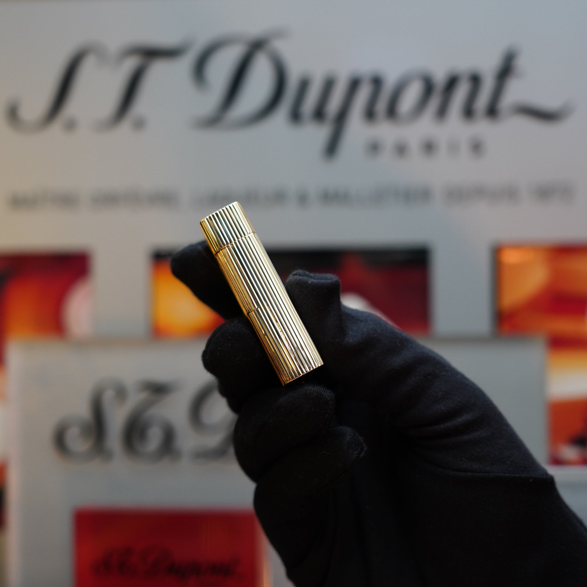 A gloved hand holding the illustrious 1960 S.T. Dupont BS lighter, crafted from 18k solid gold with a 750 hallmark, in front of a blurred S.T. Dupont advertisement background—a true collector's item.