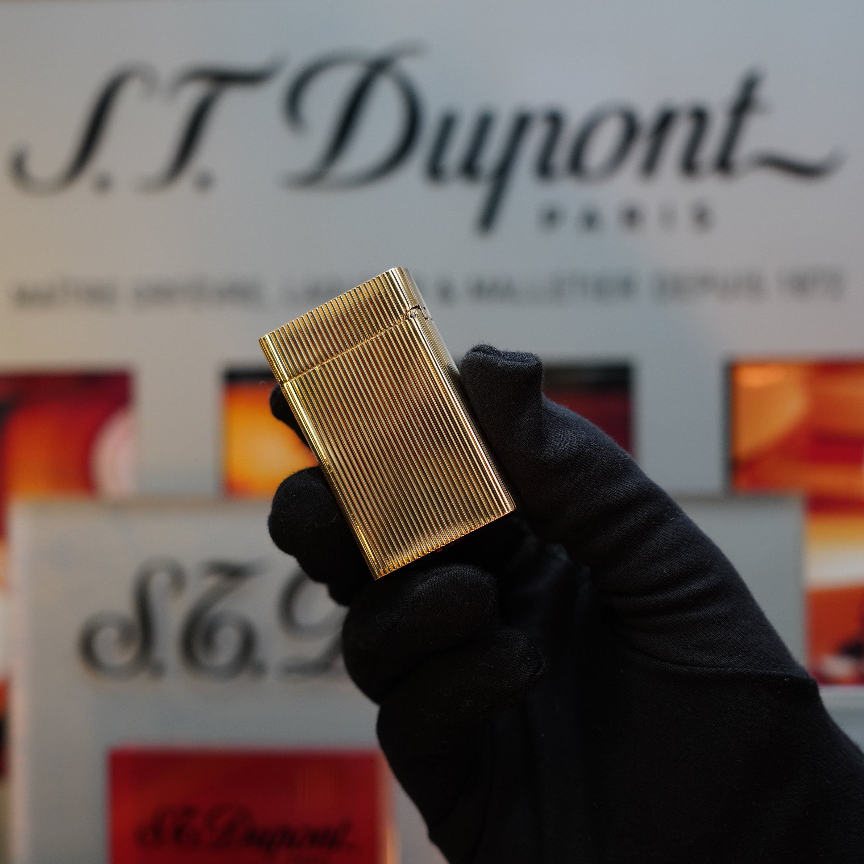 A gloved hand holds a 1960 S.T. Dupont BS 18k solid gold lighter featuring a ridged design and the 750 hallmark, a true collector's item from the 60s. The background includes blurred S.T. Dupont branding and products.