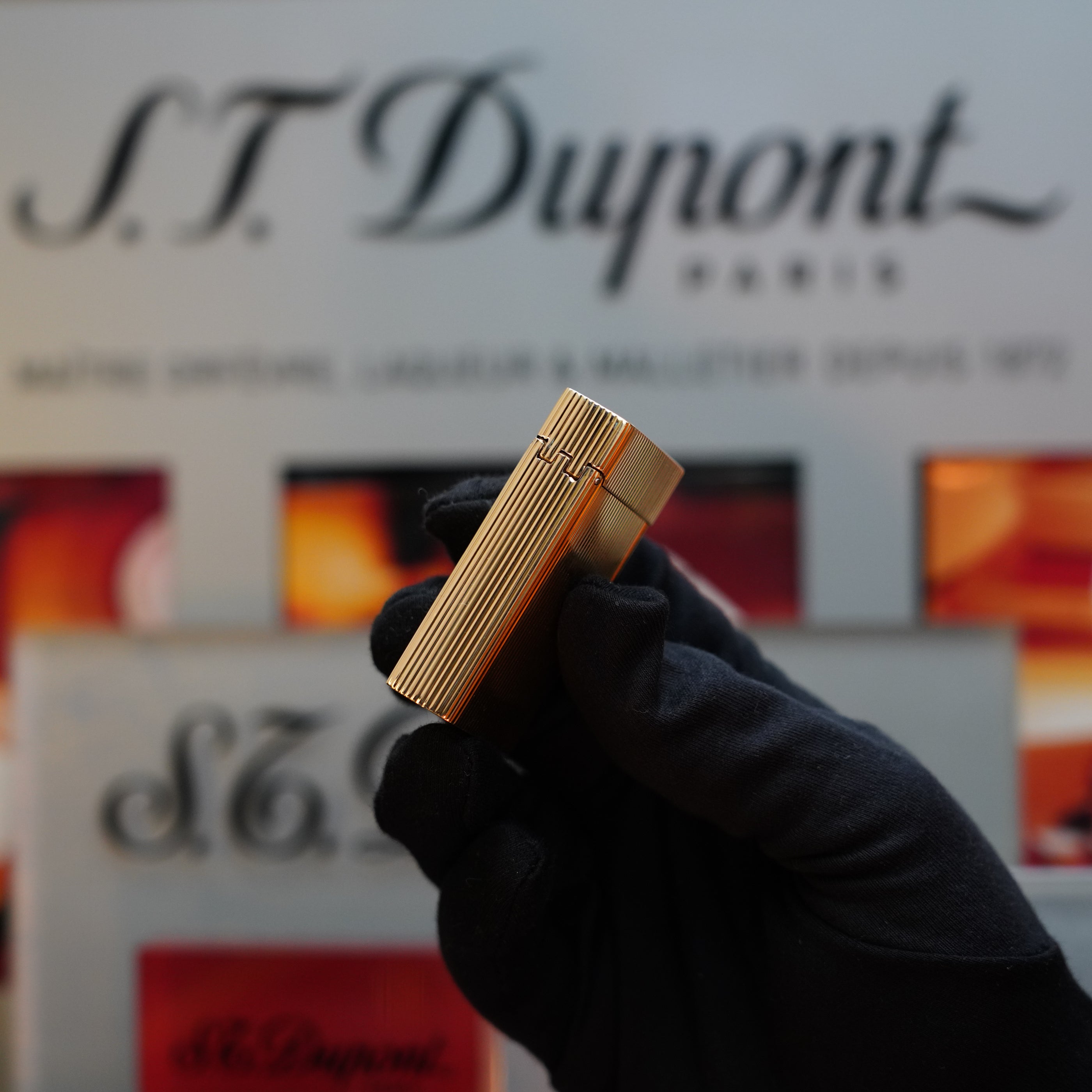 A gloved hand holding a vintage 1960 S.T. Dupont BS 18k Solid Gold Lighter with the 750 hallmark in front of a branded display background, showcasing this exquisite collector's item from the 60s.