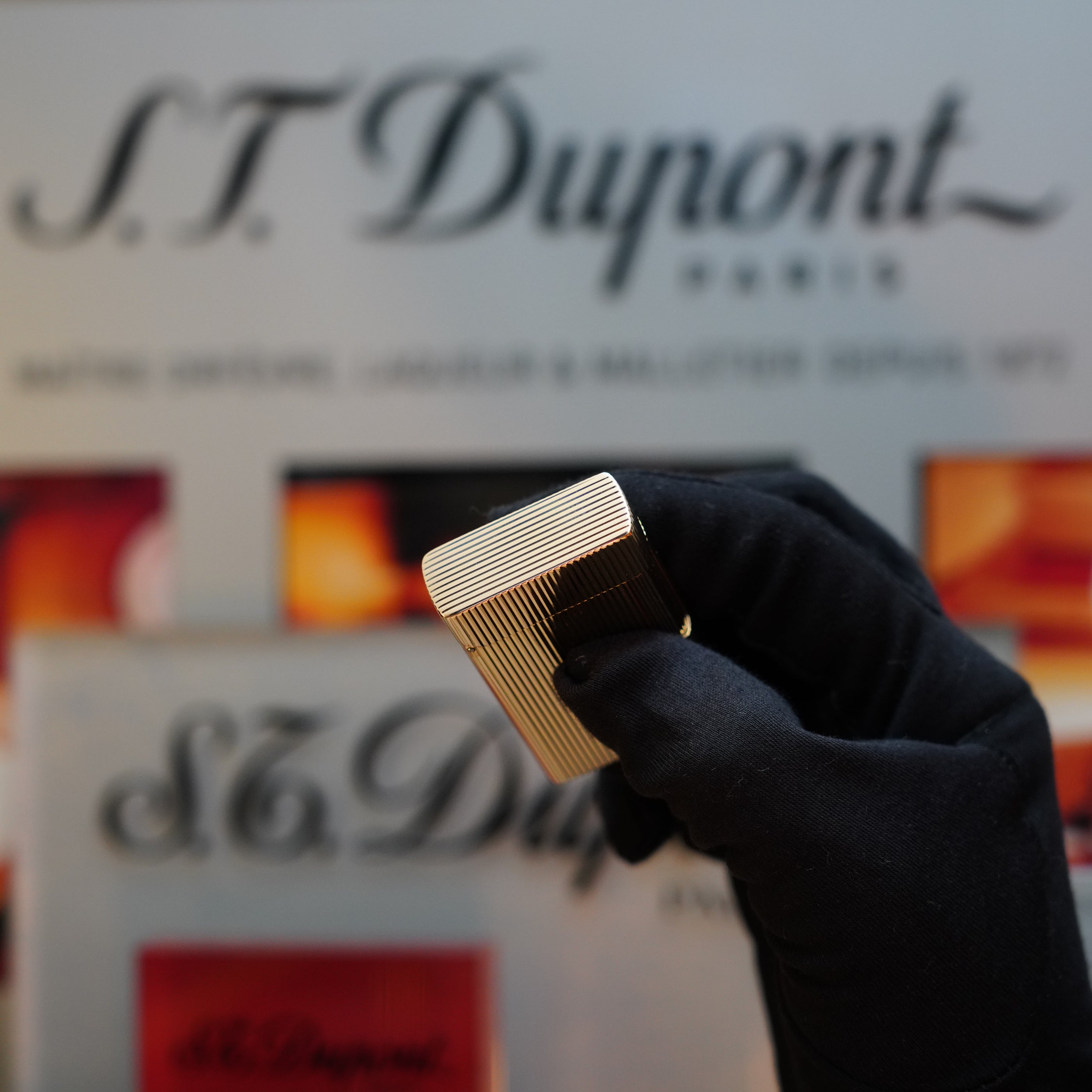 A black-gloved hand holding an exquisite 1960 St Dupont BS 18k solid gold lighter with the 750 hallmark, with a blurred S.T. Dupont Paris sign in the background. This vintage S.T. Dupont lighter is a true collector's item from the 60s.