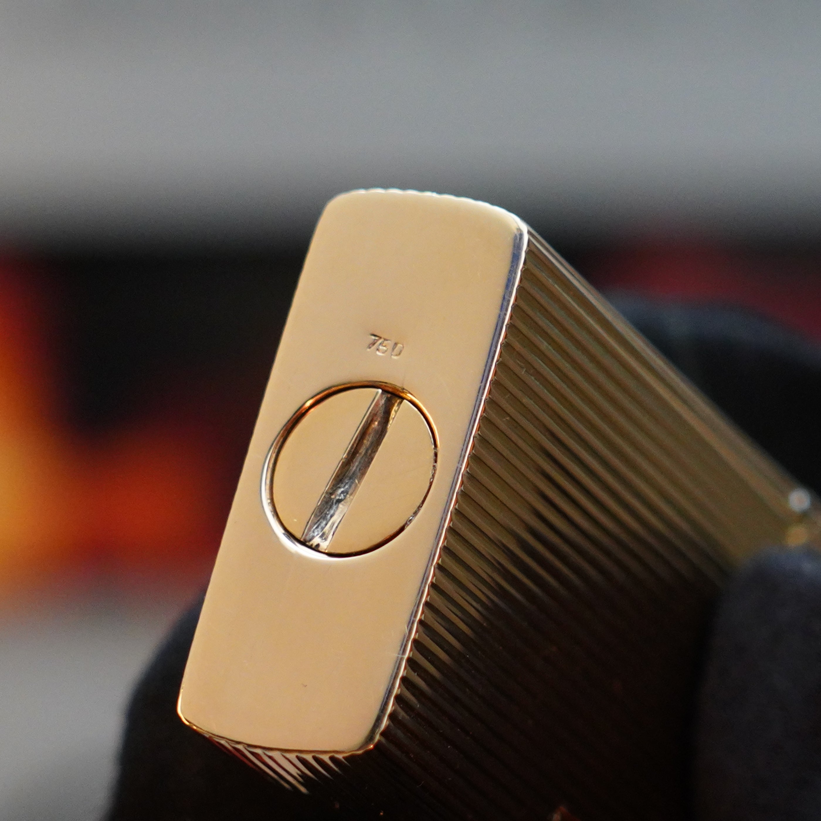 A close-up of a metallic object with a ridged surface, featuring a circular component and the engraved text "7ED." The object, reminiscent of a vintage 1960 St Dupont BS 18k Solid Gold Lighter with 750 hallmark from S.T. Dupont, is held by a hand wearing a black glove.