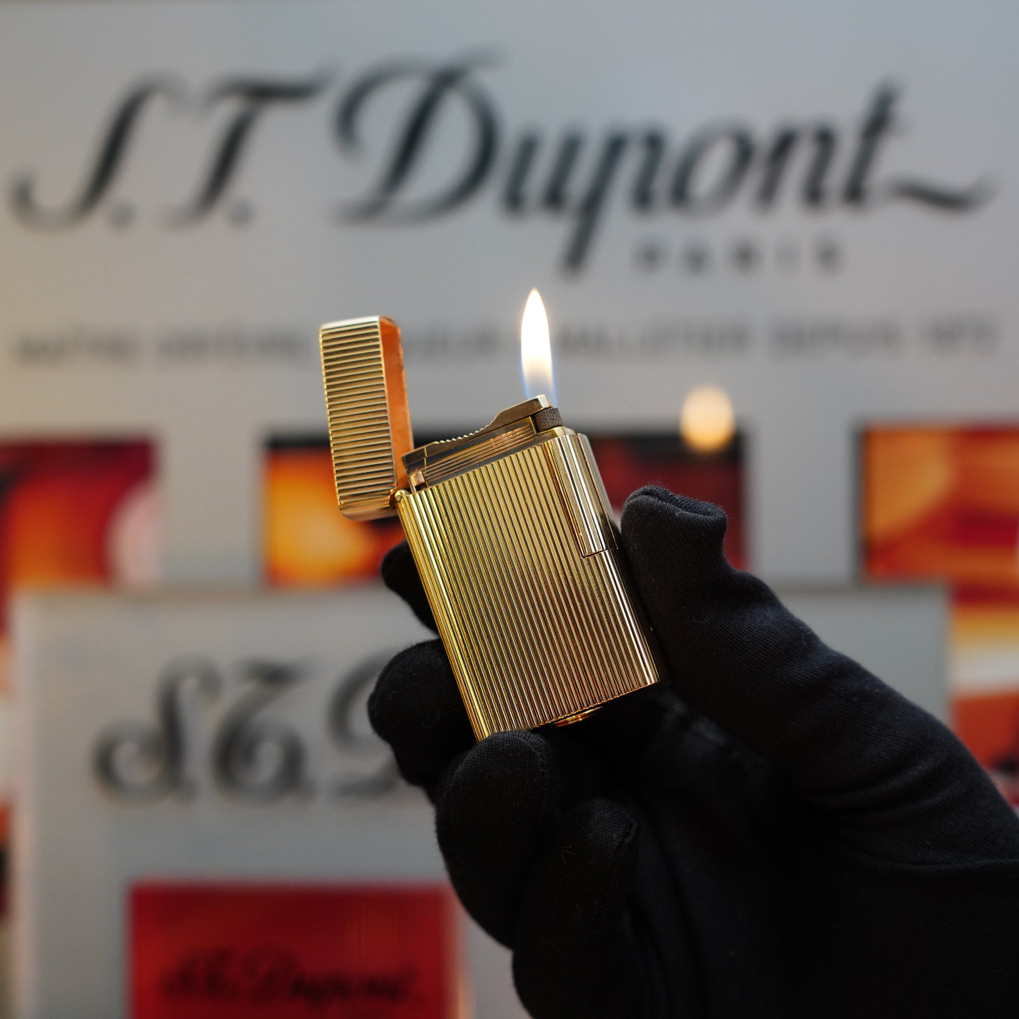 A gloved hand holds a vintage 1960 St Dupont BS 18k Solid Gold Lighter with a flame, in front of a blurred S.T. Dupont Paris sign. This exquisite 750 hallmark lighter, a true collector's item from the '60s, exudes timeless elegance and sophistication.