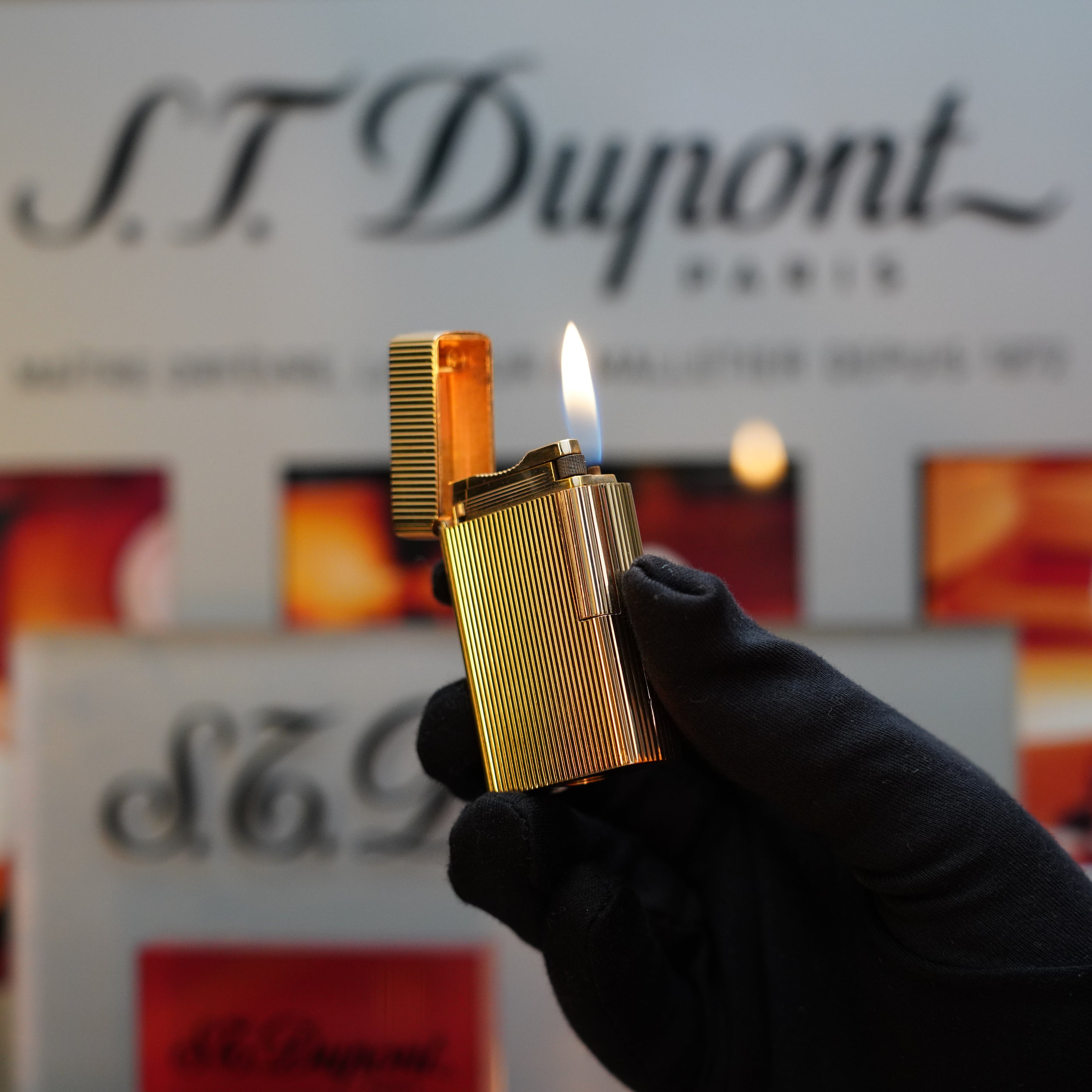 Close-up of a hand in a black glove holding an S.T. Dupont 1960 St Dupont BS 18k Solid Gold Lighter with a flame, set against a blurred background featuring S.T. Dupont branding. This collector's item from the 60s boasts elegance and craftsmanship.