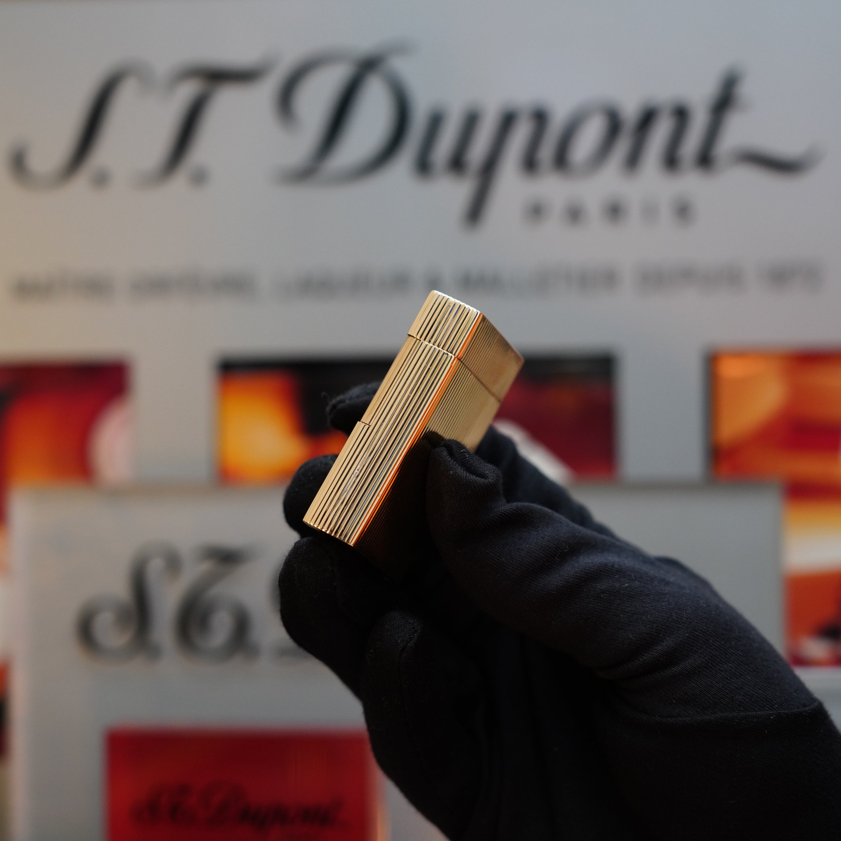 A gloved hand holding a 1960 St Dupont BS 18k Solid Gold Lighter with 750 hallmark in front of an S.T. Dupont Paris background, showcasing a classic collector's item from the iconic brand.