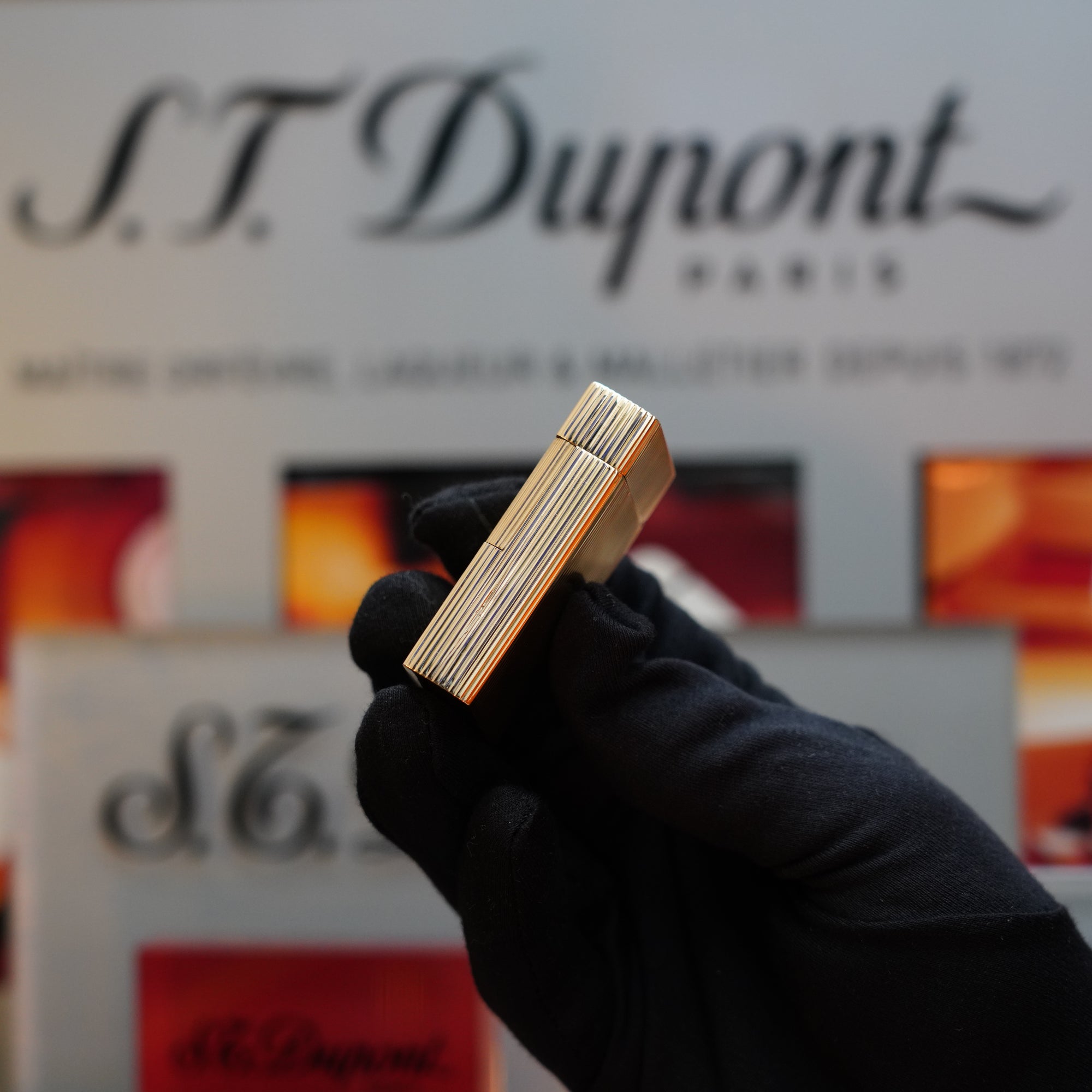 A hand in a black glove holds a 1960 St Dupont BS 18k Solid Gold Lighter with the 750 hallmark in front of a blurred background featuring the S.T. Dupont logo and branding, highlighting this collector's item from the 60s.