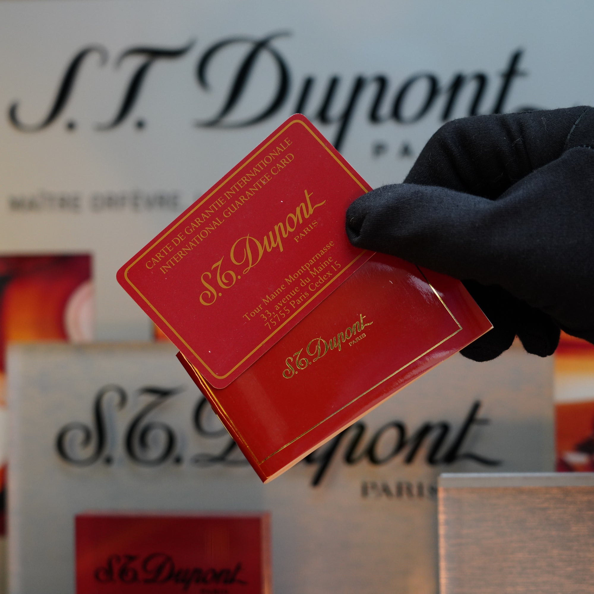 A gloved hand holding a red S.T. Dupont card with gold lettering, promoting an international guarantee. In the background, the S.T. Dupont logo and other packaging items are displayed next to a collector's item: the exquisite 1960 St Dupont BS 18k Solid Gold Lighter with 750 hallmark.