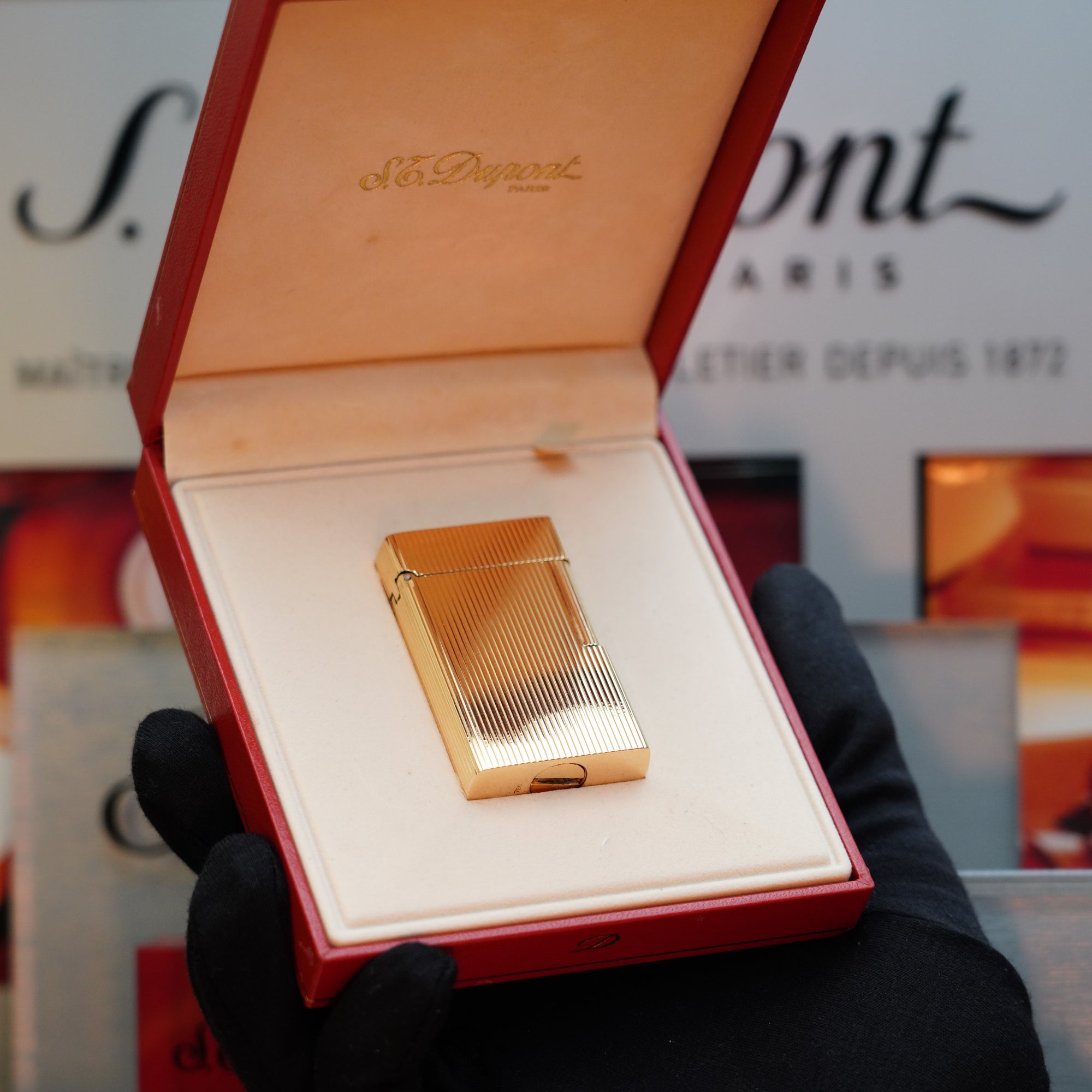 A person wearing black gloves holds an open red box containing a 1960 St Dupont BS lighter, crafted from 18k solid gold and bearing the 750 hallmark, with "S.T. Dupont" engraved on the lid. This collector's item from the brand S.T. Dupont stands out against the blurred S.T. Dupont Paris sign in the background.