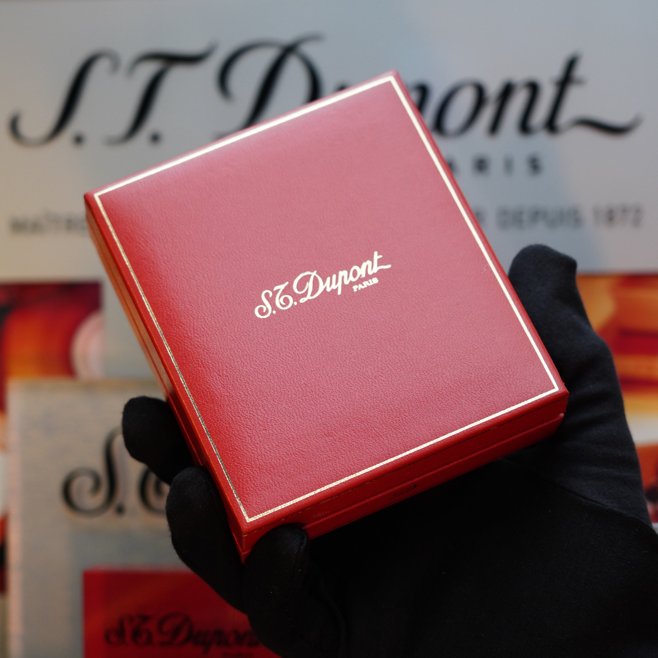 A gloved hand holds a red S.T. Dupont Paris box, adorned with white text branding on the lid, cradling a vintage 1960 St Dupont BS 18k Solid Gold Lighter with a 750 hallmark.