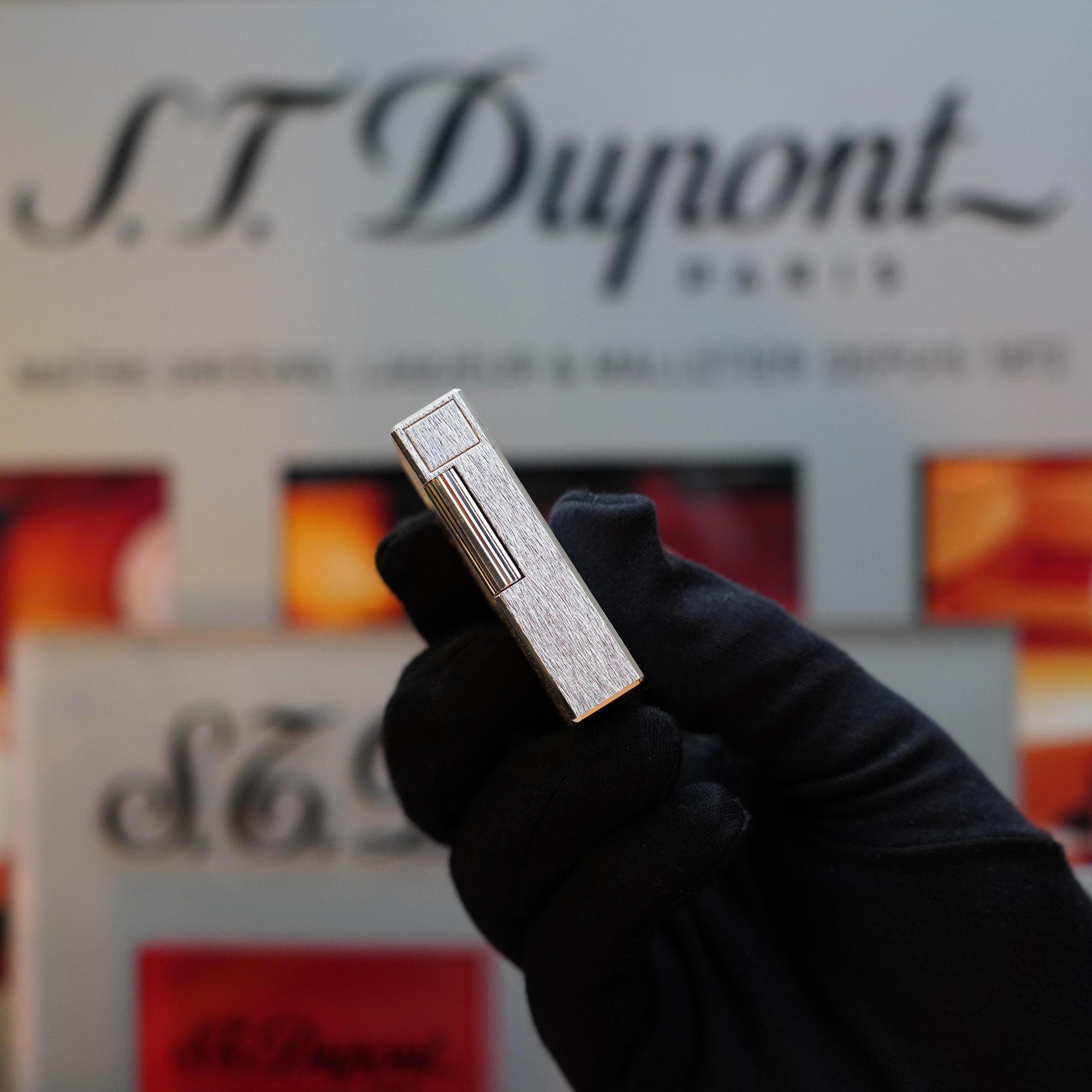 A hand in a black glove holds a 1980 St Dupont Heavy Silver Finish Ligne 1 Large Rare Pattern lighter. Blurred S.T. Dupont branding and red-orange images are in the background, highlighting this collector's item.