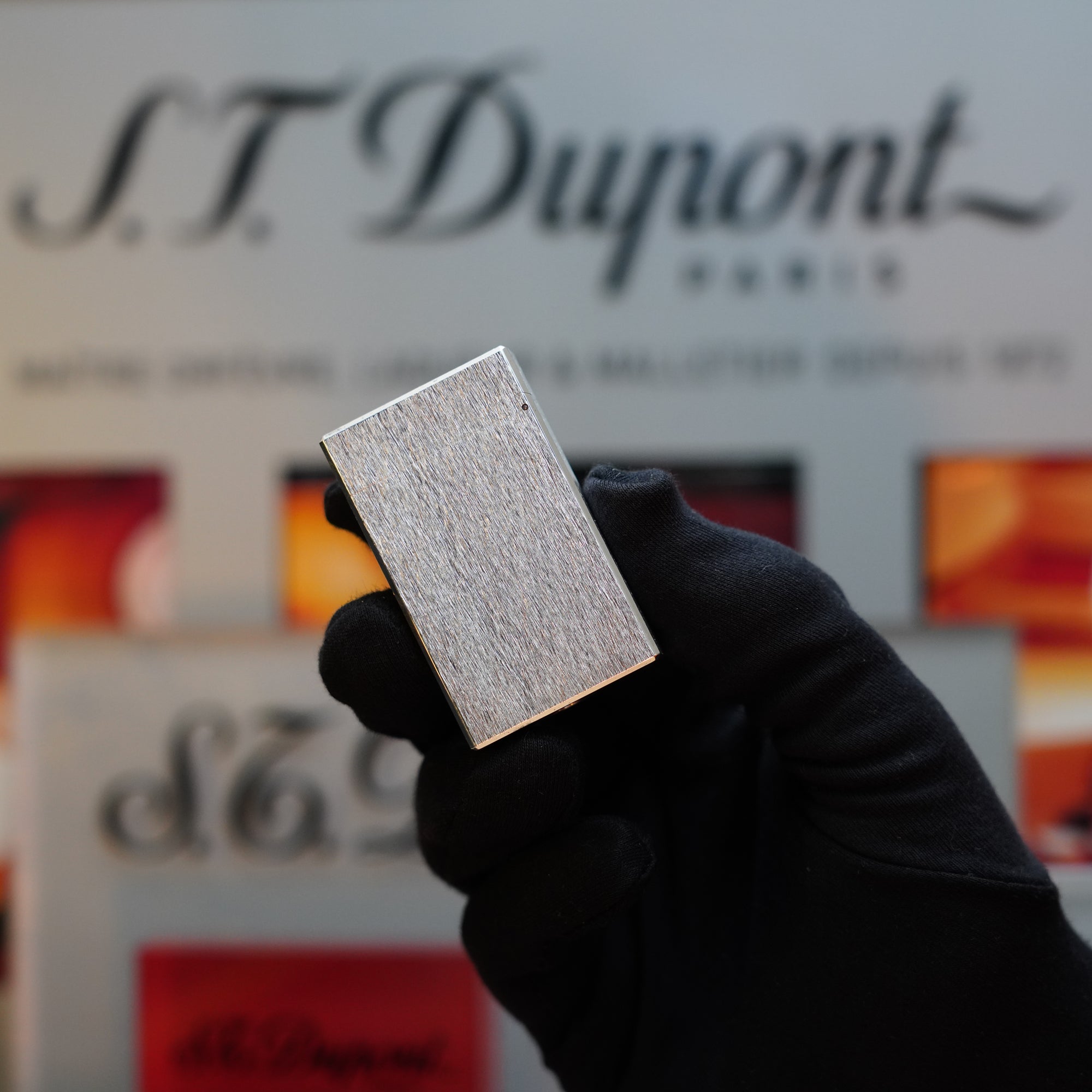 A gloved hand holds up a 1980 S.T. Dupont Heavy Silver Finish Ligne 1 Large Lighter with a rare pattern in front of a blurred background displaying the S.T. Dupont logo and various lighter designs, making this collector's item stand out beautifully.