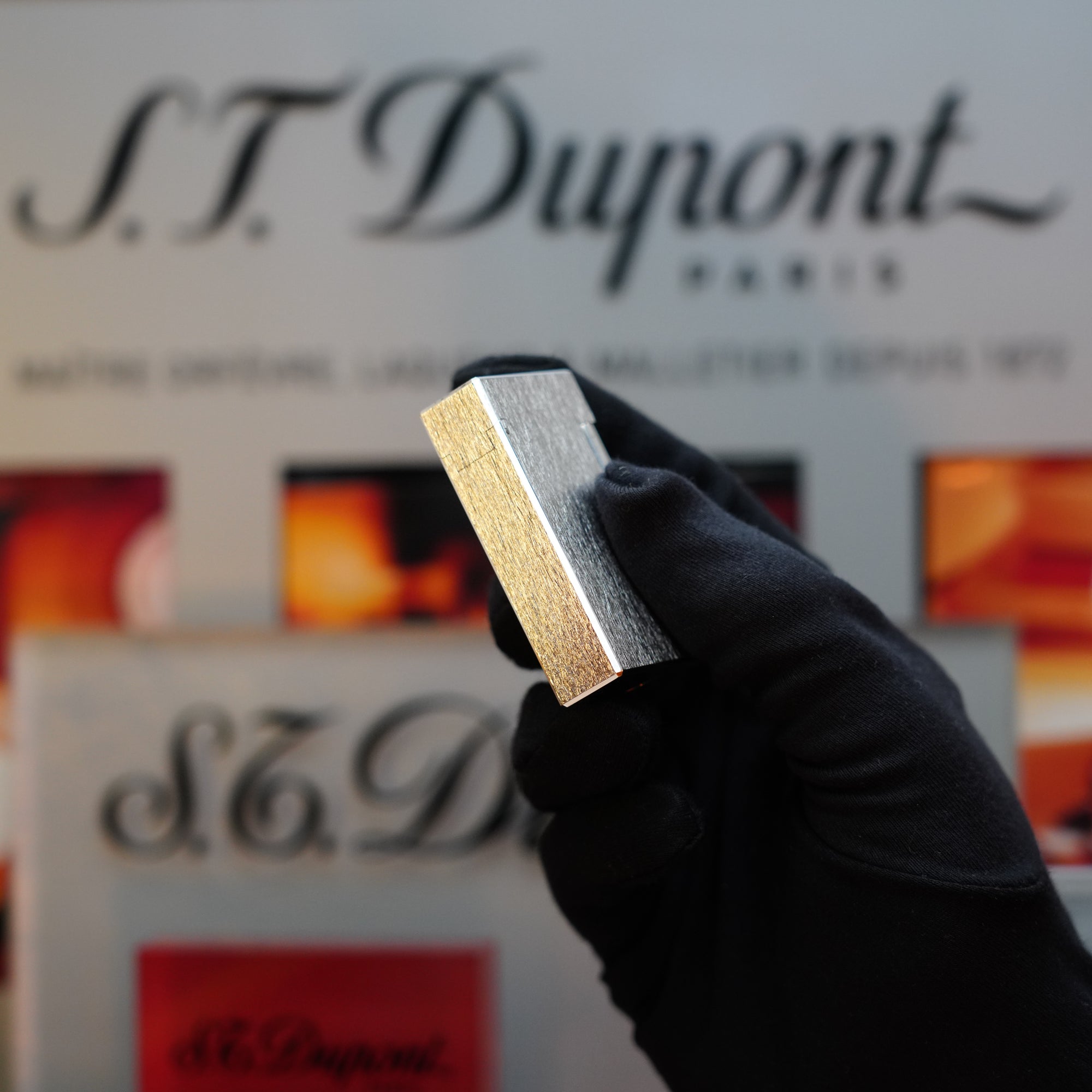 A gloved hand holds a silver rectangular object, a 1980 St Dupont Heavy Silver Finish Ligne 1 Large Rare Pattern lighter, in front of a blurred S.T. Dupont Paris sign.