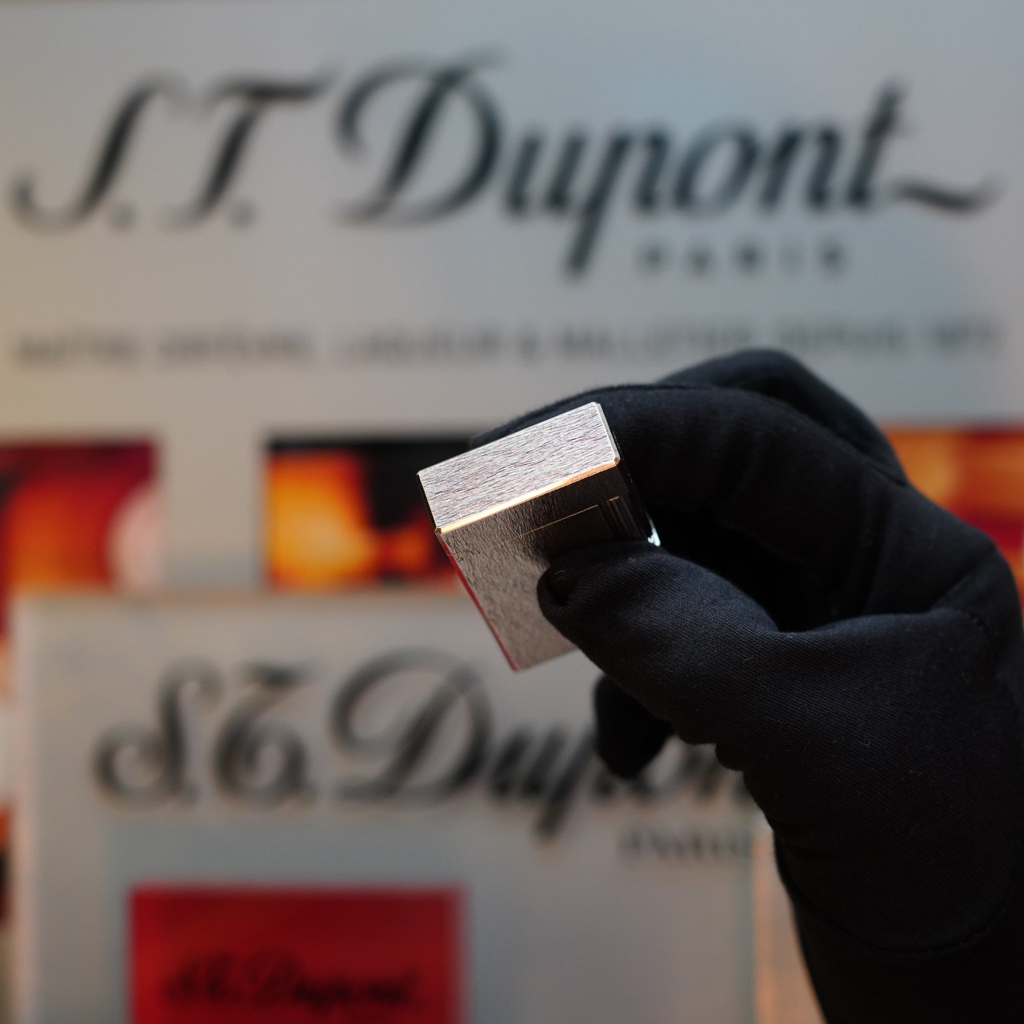 A gloved hand holds a 1980 St Dupont Heavy Silver Finish Ligne 1 Large Rare Pattern lighter in front of promotional signage for the S.T. Dupont brand.