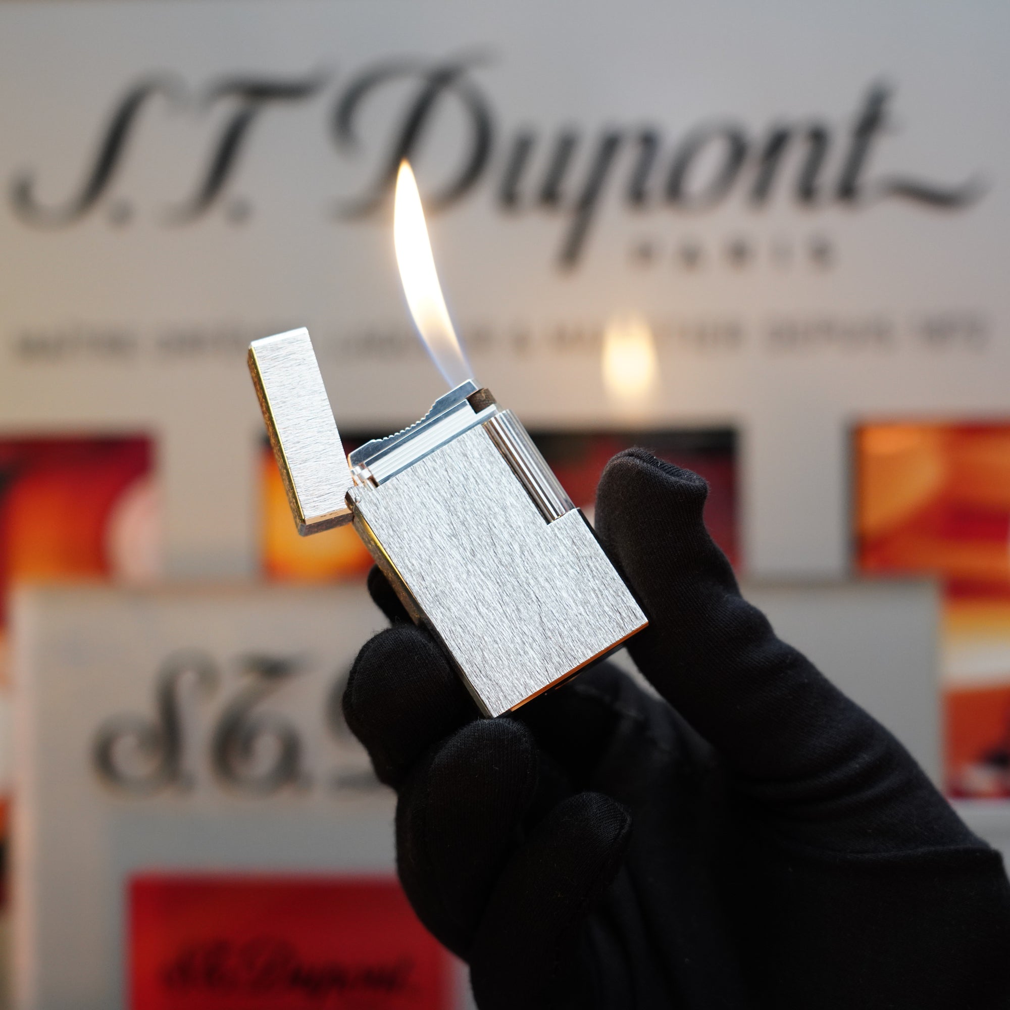 A gloved hand holds a lit 1980 St Dupont Heavy Silver Finish Ligne 1 Large Rare Pattern lighter. The iconic S.T. Dupont branding is visible in the background, marking it as a true collector's item.