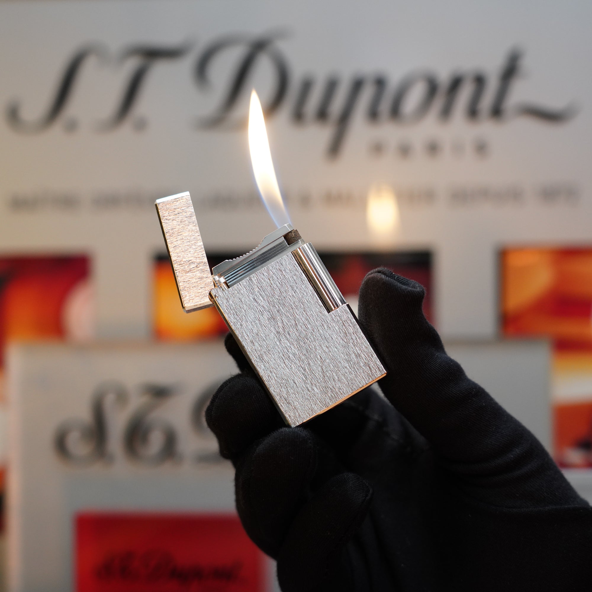 A gloved hand holding a lit "1980 St Dupont Heavy Silver Finish Ligne 1 Large Rare Pattern" lighter in front of a blurred background with "S.T. Dupont Paris" text, highlighting this collector's item.