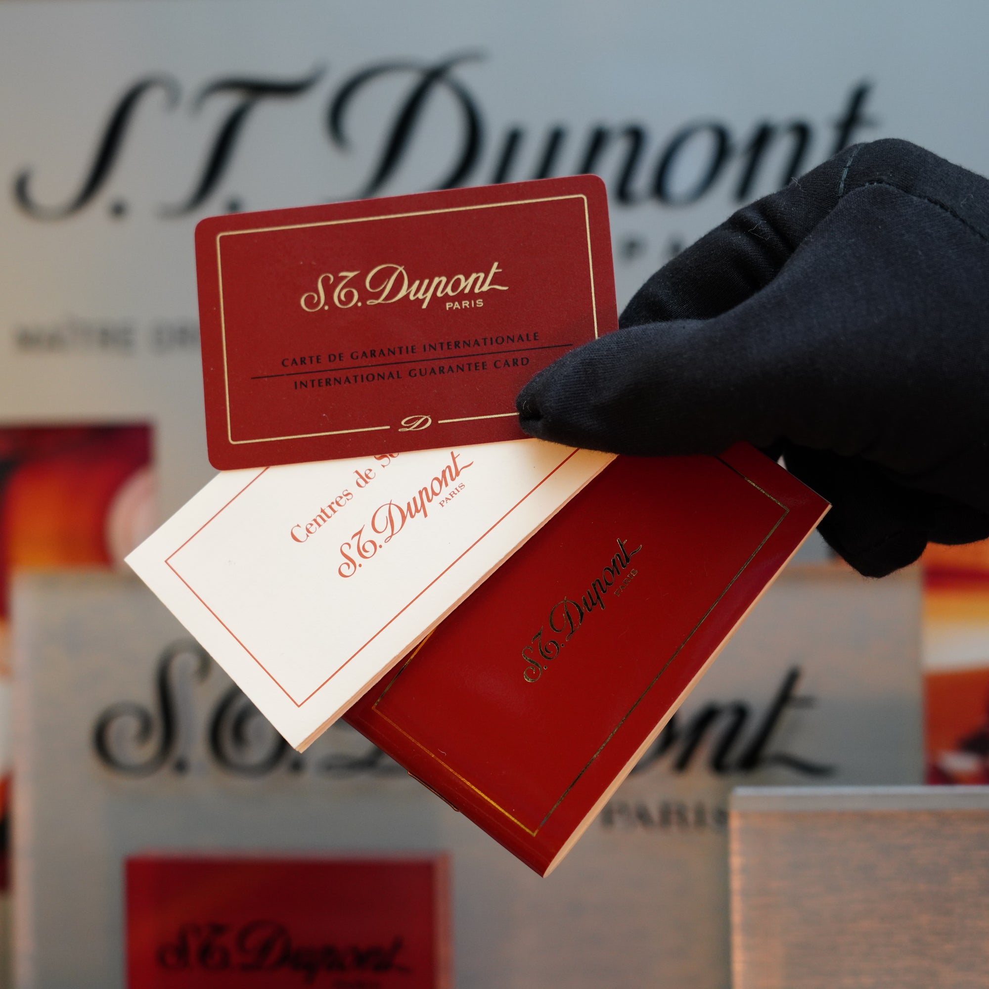 A hand in a black glove holds two S.T. Dupont International Guarantee Cards, one red and one white, in front of a display of S.T. Dupont branded items, including the coveted 1980 St Dupont Heavy Silver Finish Ligne 1 Large Rare Pattern—a true collector's item for enthusiasts.