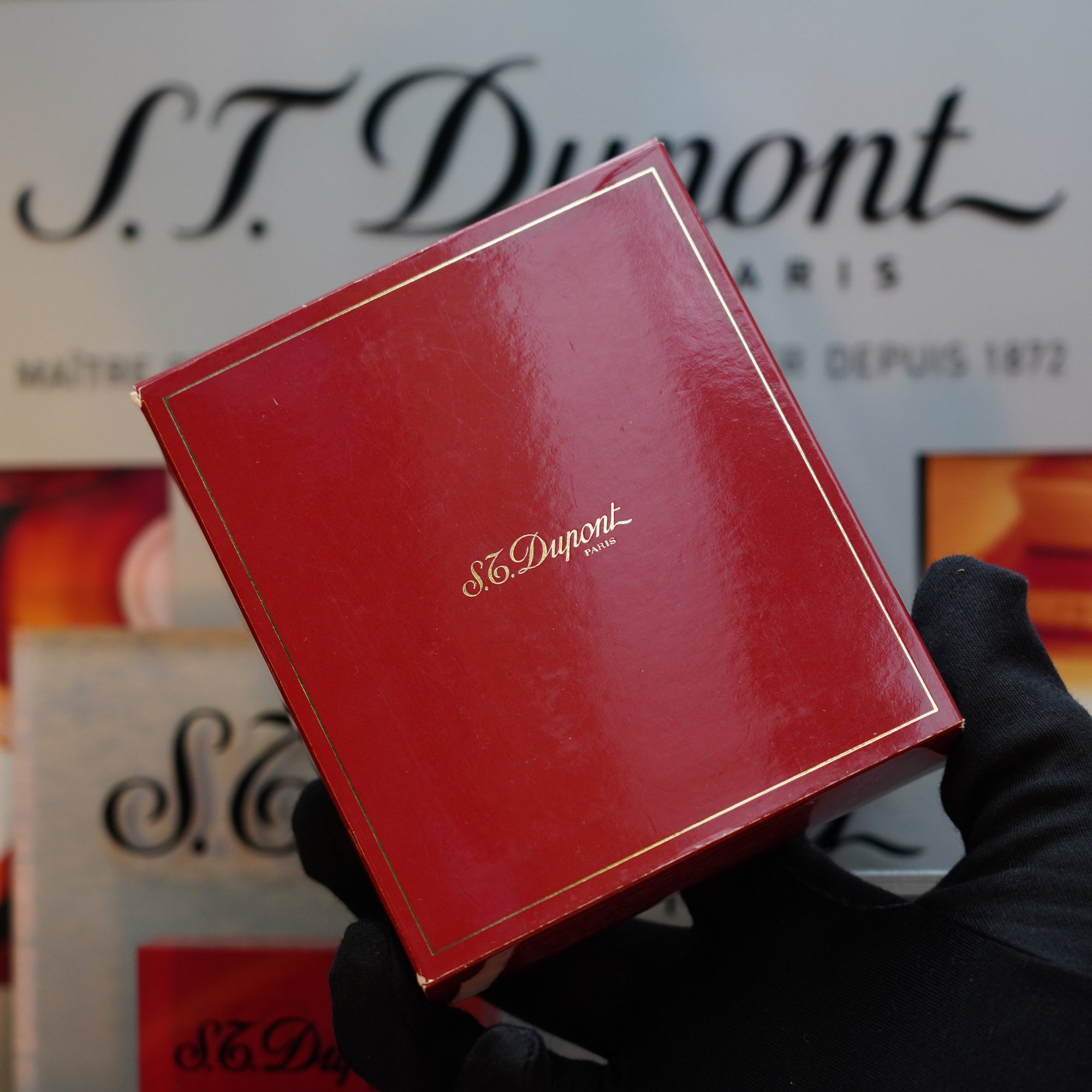 A gloved hand holds a red S.T. Dupont Paris box in front of a blurred background displaying the brand's logo and text, showcasing a 1980 St Dupont Heavy Silver Finish Ligne 1 Large Rare Pattern lighter.