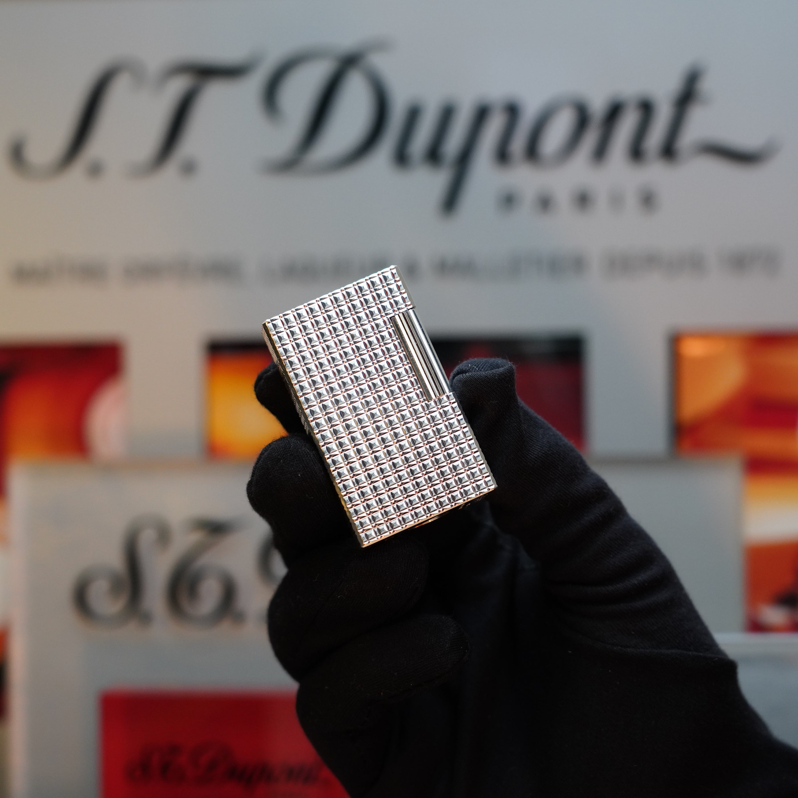 A gloved hand holds a checkered metal lighter, the 1980 S.T. Dupont Silver Finish Rare Pattern Large Size Ligne 1 Lighter, in front of a blurred background featuring the S.T. Dupont Paris logo—a true collector's item.