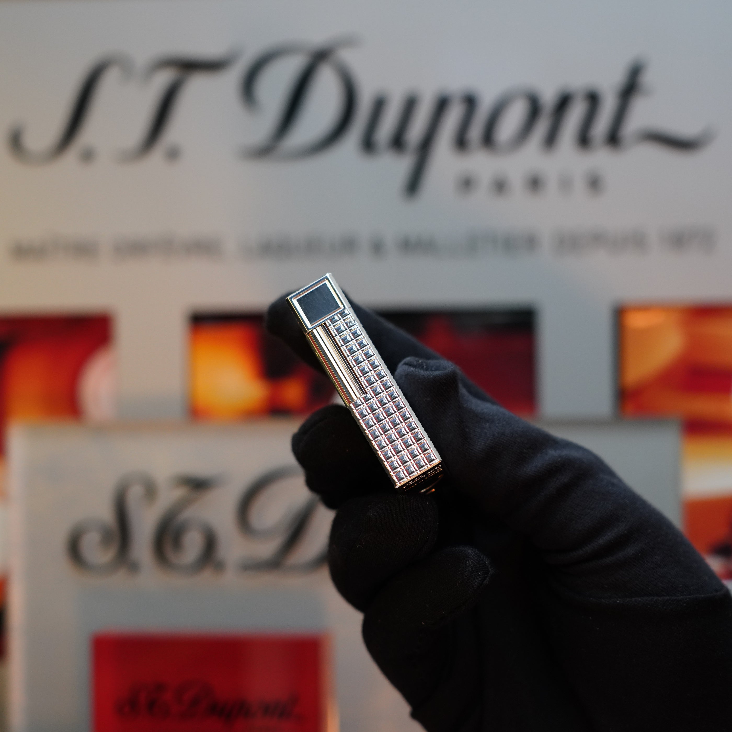 A gloved hand holds a 1980 S.T. Dupont Silver Finish Rare Pattern Large Size Ligne 1 Lighter against a blurred background featuring the iconic S.T. Dupont logo and text, making it a true collector's item.