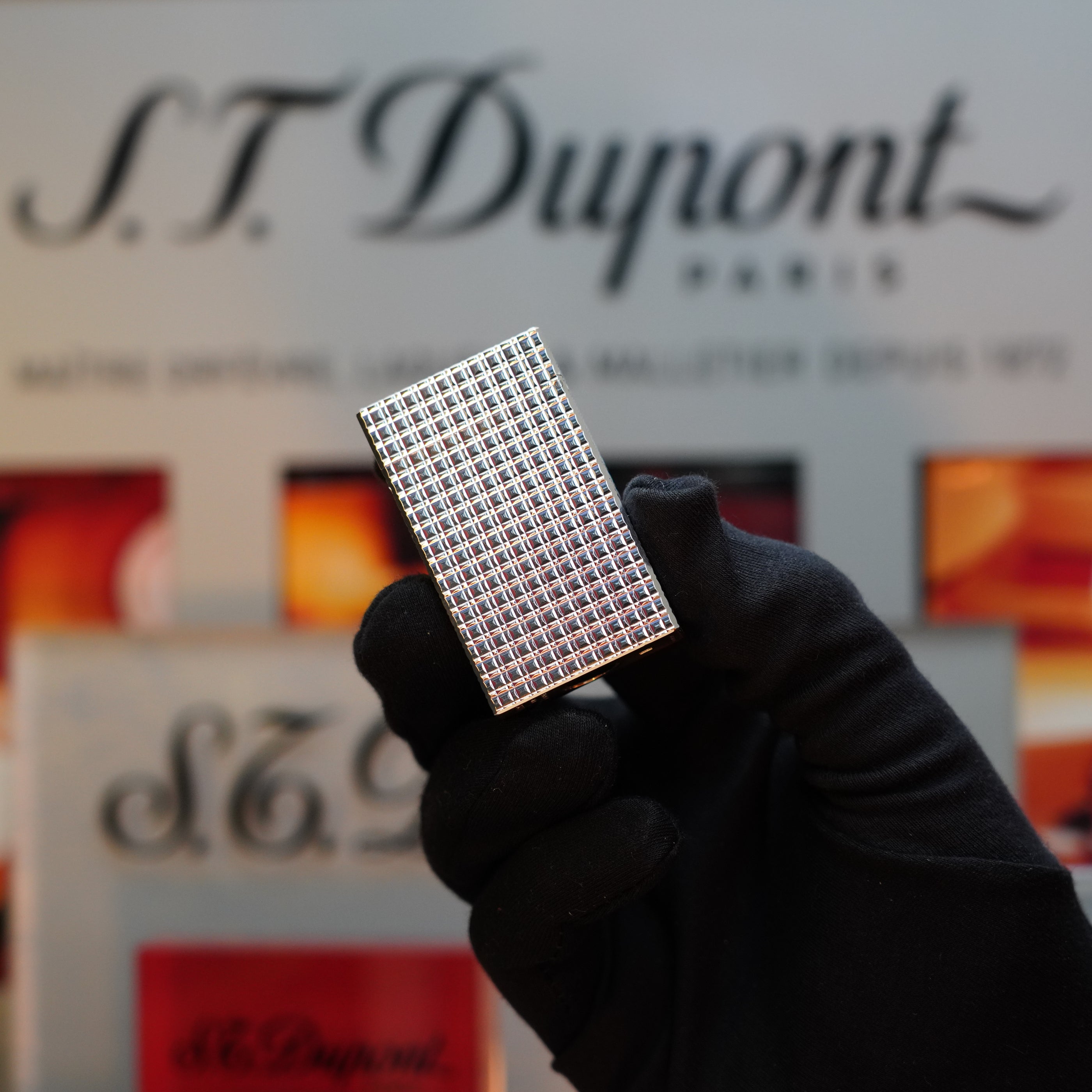 A gloved hand holds a silver checkered rectangular item—an exquisite 1980 St Dupont Silver Finish Rare Pattern Large Size Ligne 1 Lighter—in front of a blurred S.T. Dupont Paris sign.