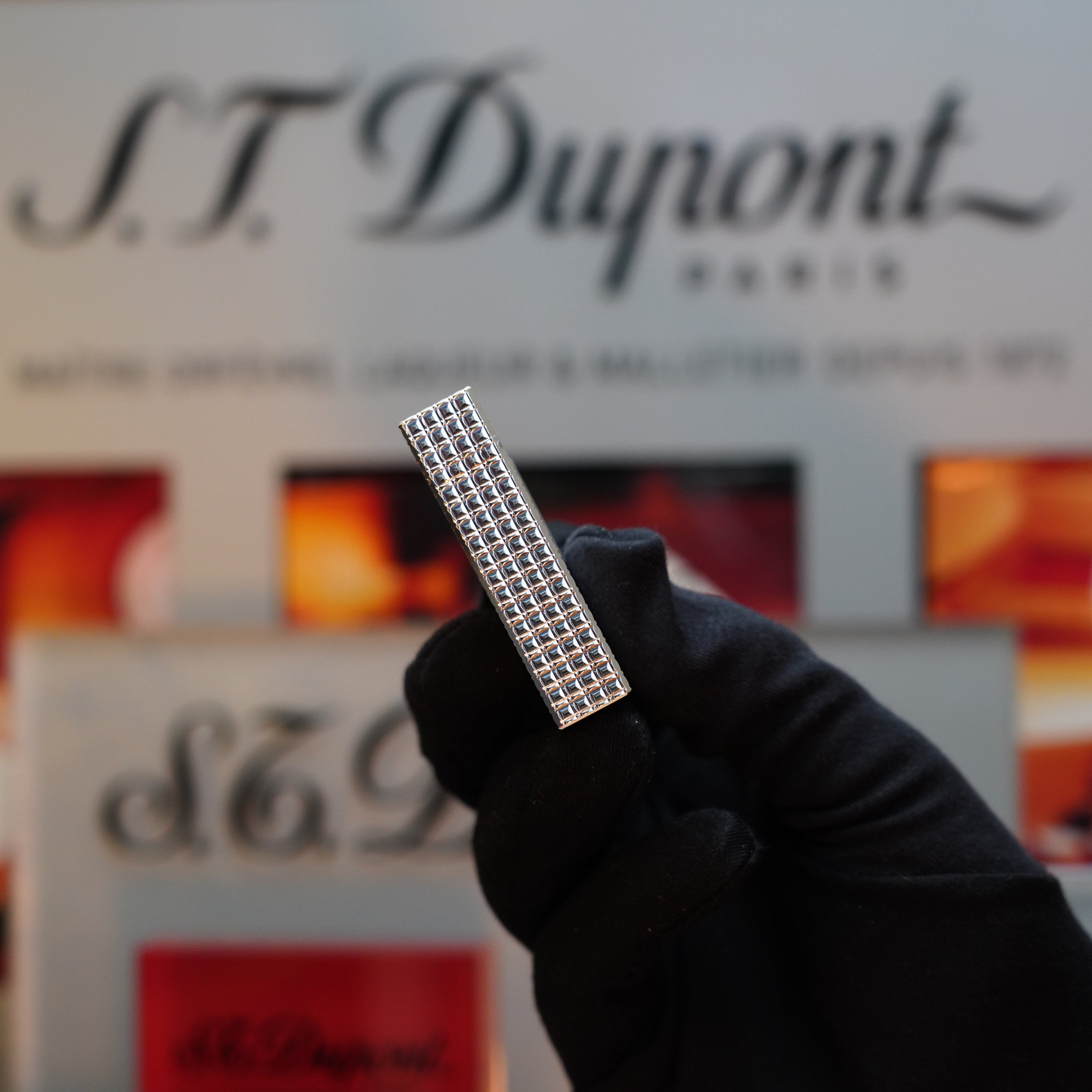 A gloved hand holds a rectangular, silver-colored grid-patterned object against a blurred background featuring the S.T. Dupont logo, showcasing the 1980 S.T. Dupont Silver Finish Rare Pattern Large Size Ligne 1 Lighter, truly a collector's item.