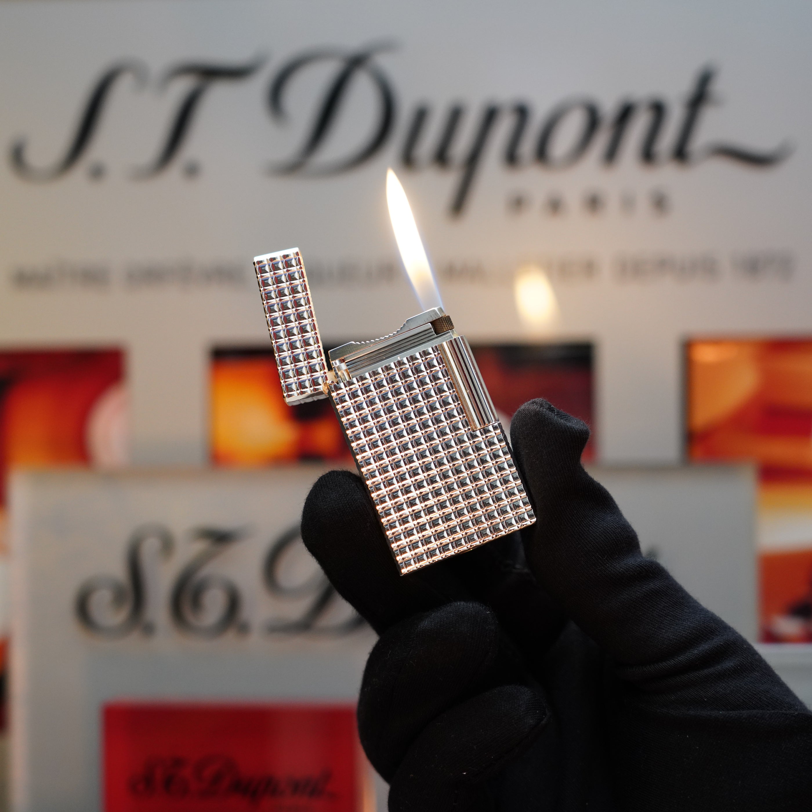 A gloved hand holds a lit 1980 S.T. Dupont Silver Finish Rare Pattern Large Size Ligne 1 lighter against a blurred background featuring the S.T. Dupont logo and promotional materials, showcasing this elegant vintage lighter as a coveted collector's item.