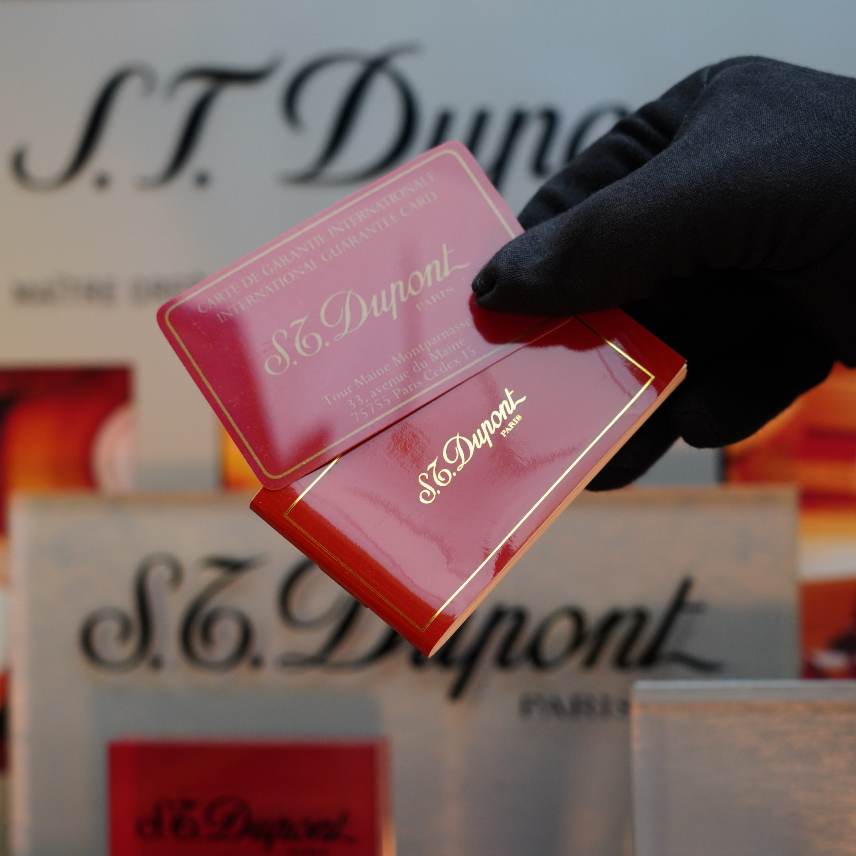 A gloved hand holds a red S.T. Dupont box with gold lettering, set against a backdrop featuring the brand's logo. Inside, discover the 1980 S.T. Dupont Silver Finish Rare Pattern Large Size Ligne 1 Lighter, a true collector's item.