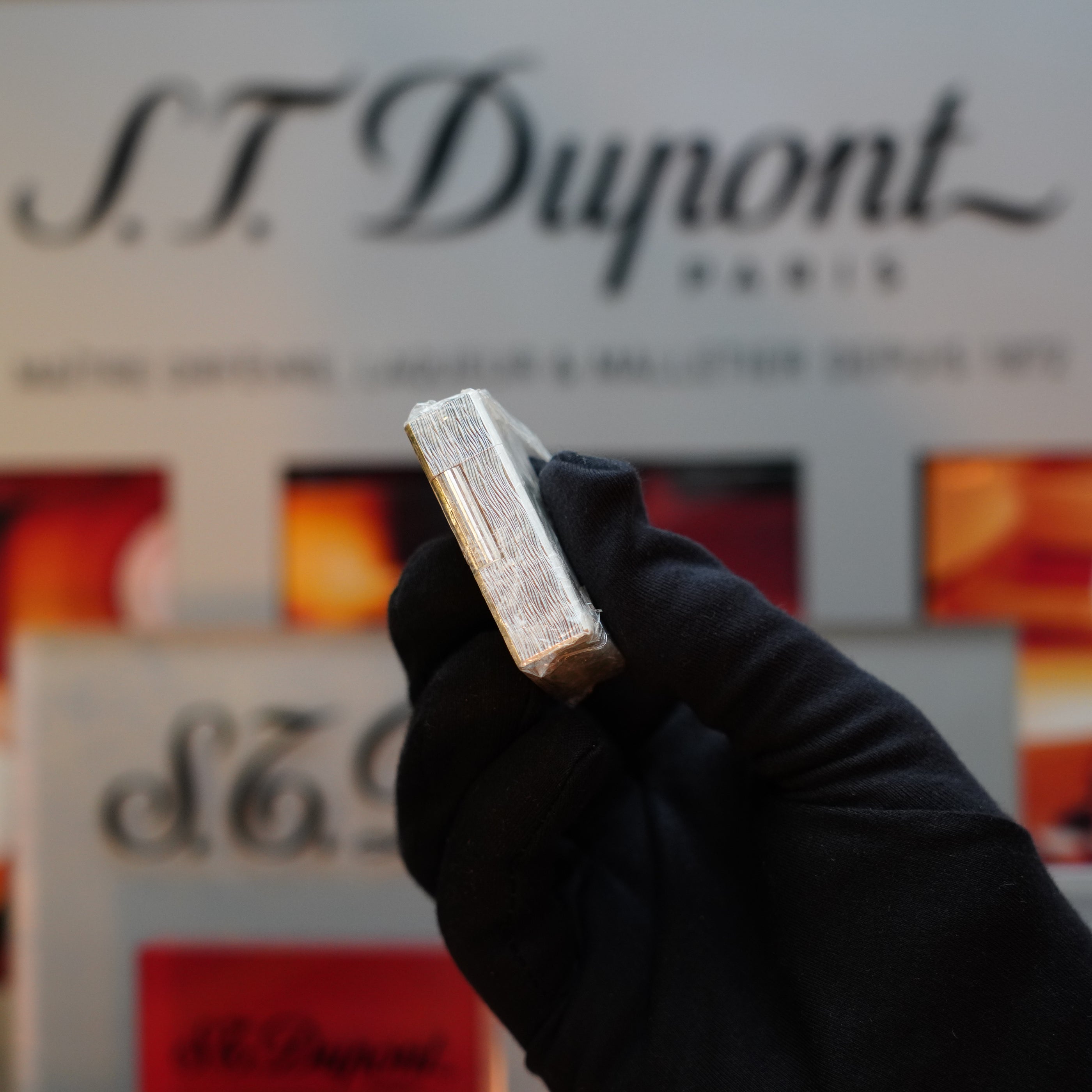 A gloved hand holding a silver metallic object with the S.T. Dupont Paris logo visible in the background, showcasing the prized 1980 St Dupont Heavy Silver Finish Ligne 1 Sealed Lighter, a coveted collector's item from the 1980s.