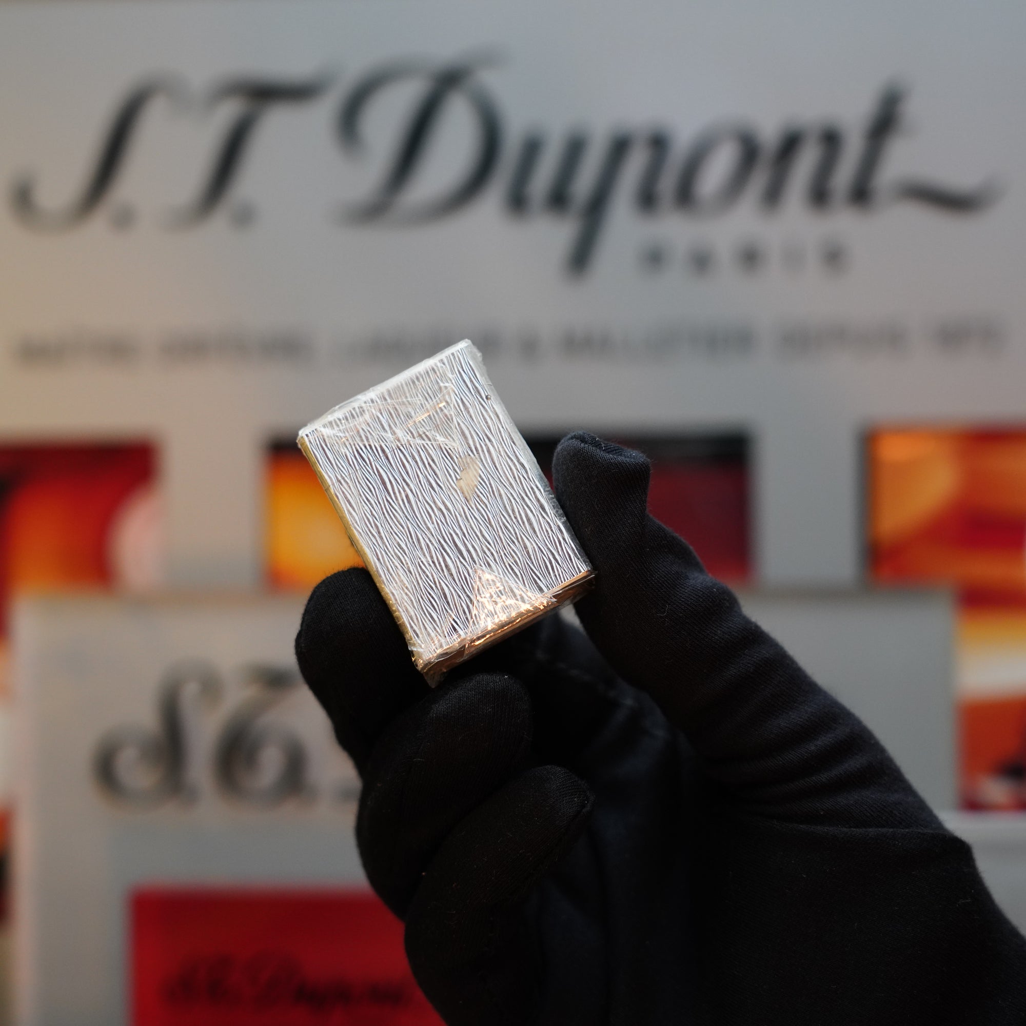 A gloved hand holding a shiny, silver metallic object—likely an S.T. Dupont 1980 Heavy Silver Finish Ligne 1 Sealed Lighter—in front of a blurred background featuring the S.T. Dupont logo.