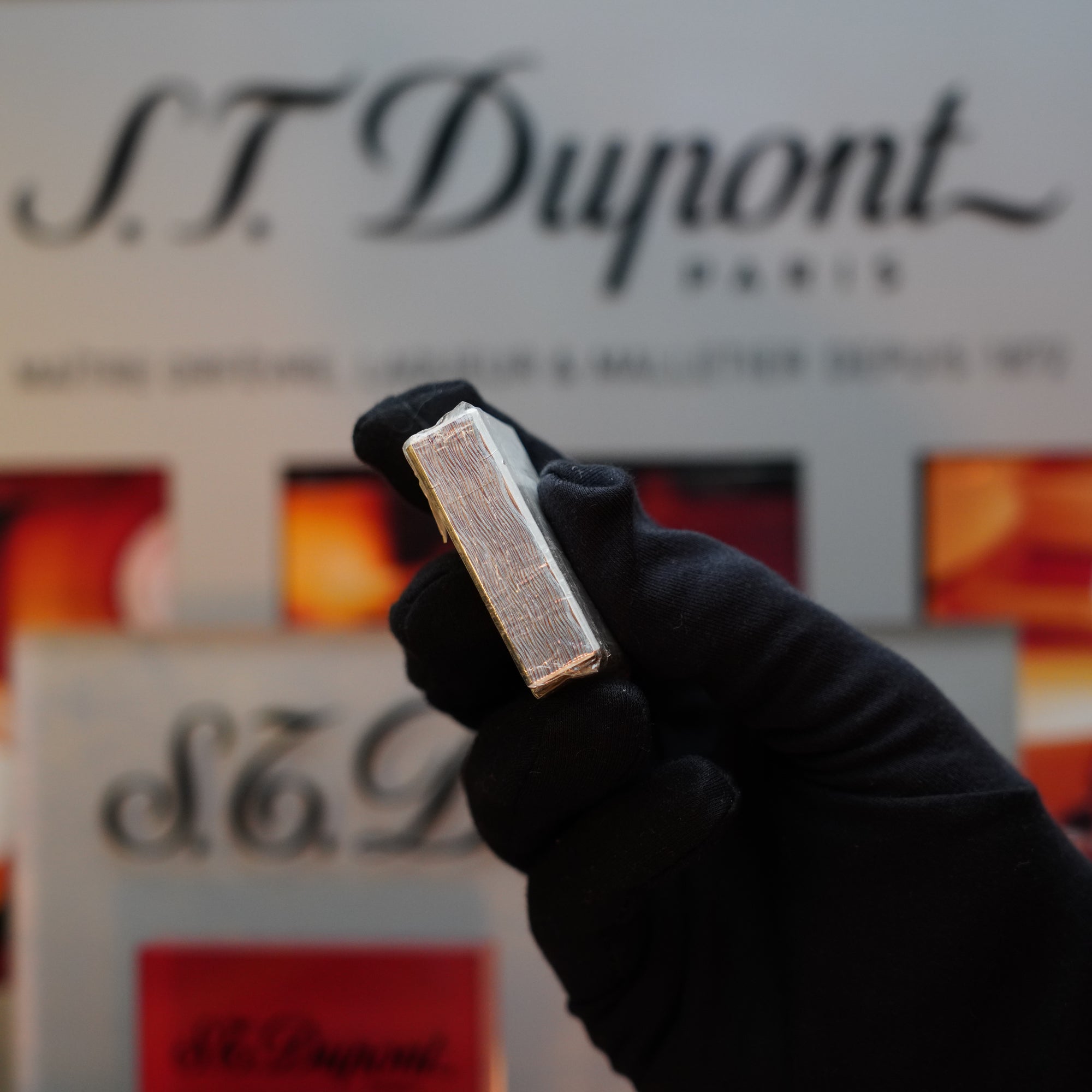 A gloved hand holds a vintage 1980 St Dupont Heavy Silver Finish Ligne 1 Sealed Lighter against a blurred background featuring the S.T. Dupont logo and various packaging, capturing the essence of this collector's item.