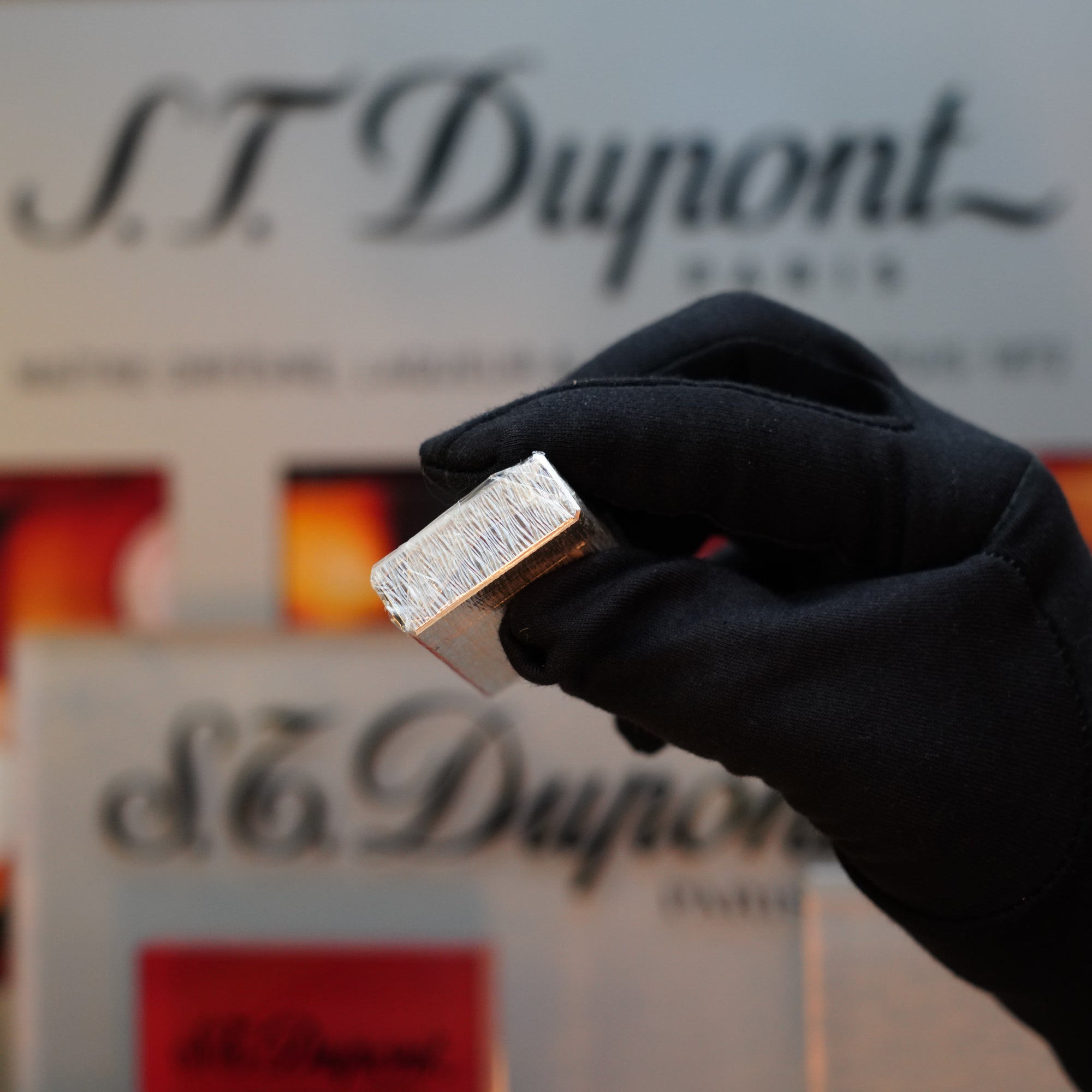 A gloved hand holding a 1980 St Dupont Heavy Silver Finish Ligne 1 Sealed Lighter, with "S.T. Dupont" branding visible in the background, highlights this classic piece as a sought-after collector's item from the 1980s.