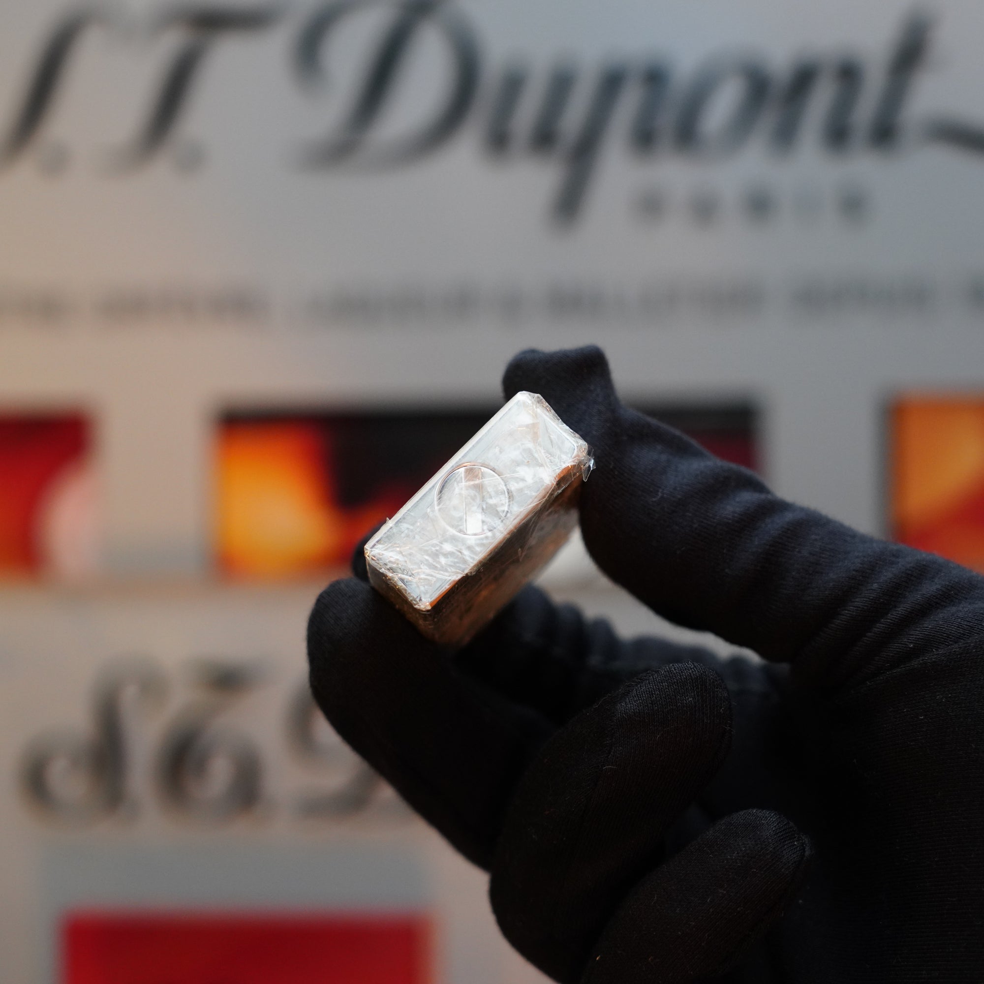 A gloved hand holds a small, silver rectangular object, likely a 1980 St Dupont Heavy Silver Finish Ligne 1 Sealed Lighter from S.T. Dupont, in front of a blurred background featuring text and red, orange, and yellow elements.