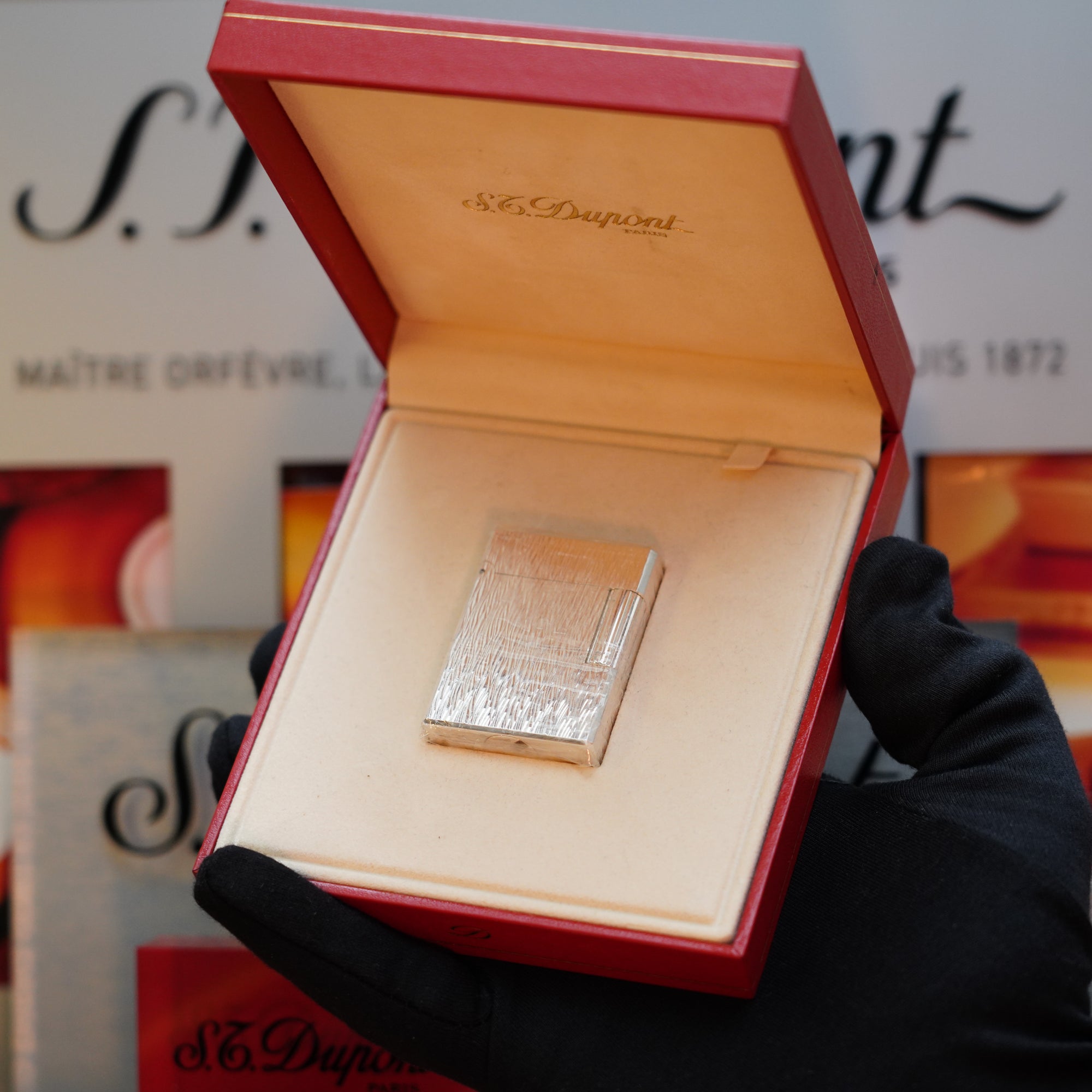 A gloved hand holds an open red box displaying a silver item, identified as a 1980 St Dupont Heavy Silver Finish Ligne 1 Sealed Lighter, with "S.T. Dupont" inscribed on the inside lid and background materials featuring the same brand name.