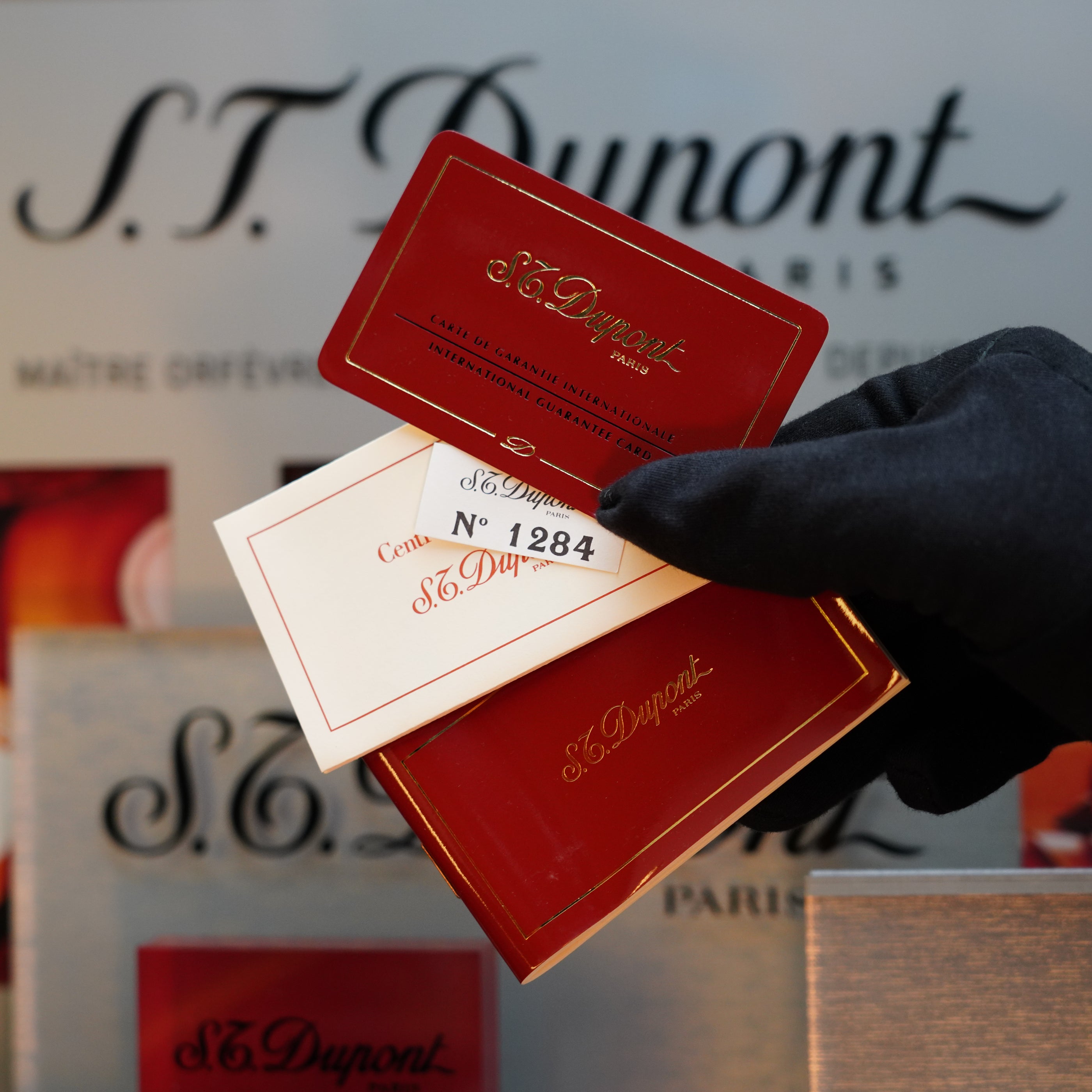 A gloved hand holds two S.T. Dupont items: a red card and a small white paper with "Cert. No 1284." The S.T. Dupont branding is visible in the background, hinting at the elegance of the 1980 St Dupont Heavy Silver Finish Ligne 1 Sealed Lighter—a true collector's item from the 1980s.