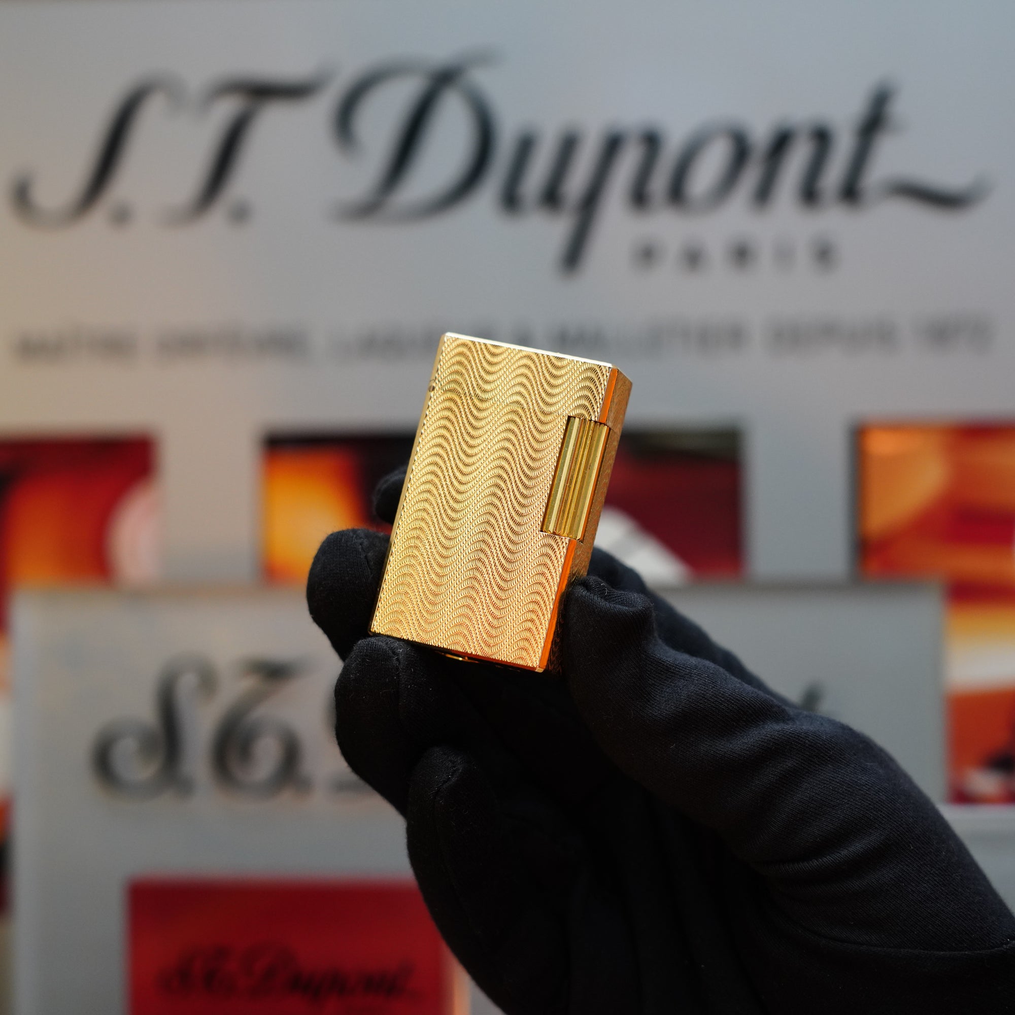 Close-up of a gloved hand holding a 1960 HERMES Lighter St Dupont Gold Finish showcasing a stunning Guilloché pattern, against a blurred background with S.T. Dupont branding and text.